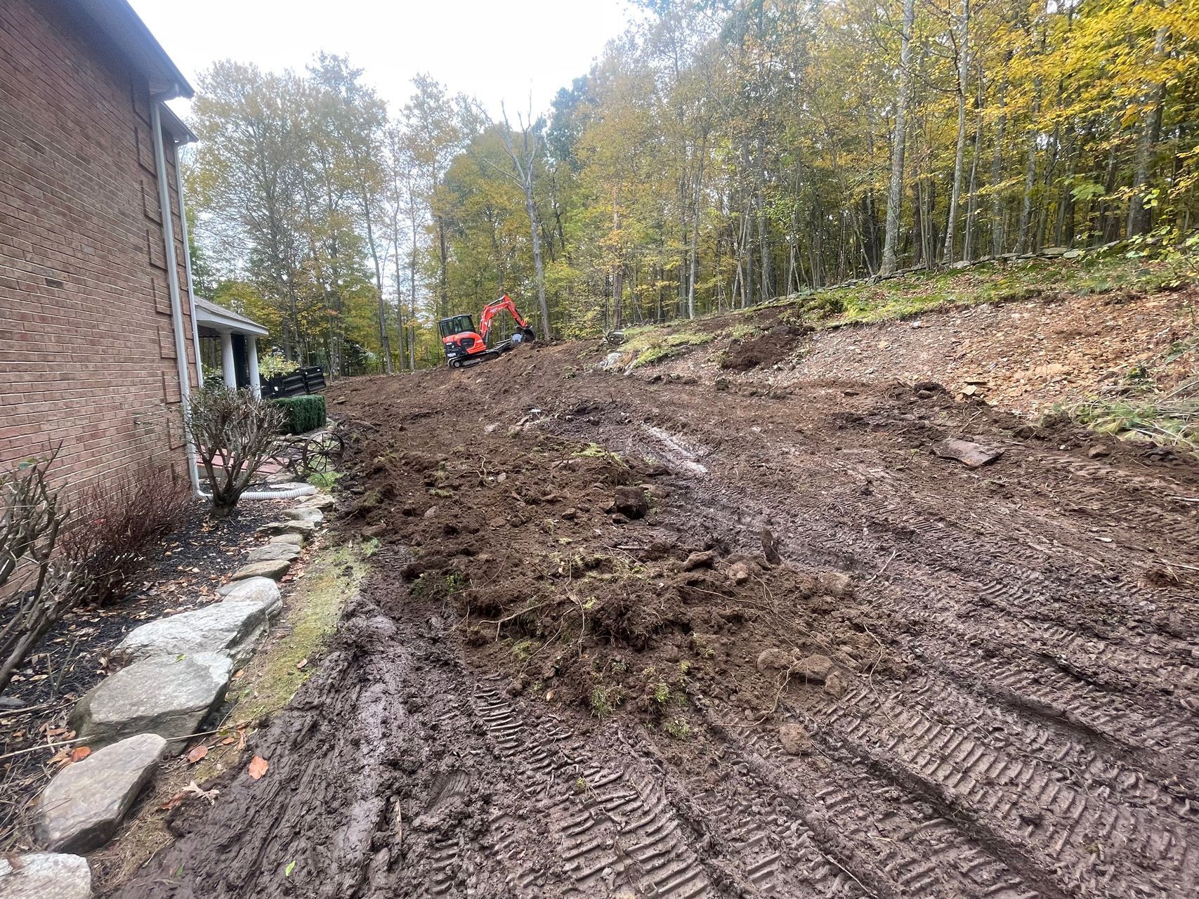 Mud-covered hillside beside a brick house, with an orange excavator and trees in the background.