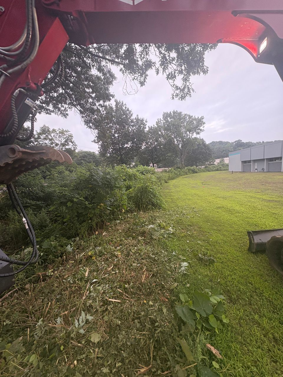 Person using a red mower to cut grass on a rainy lawn beside a gray building