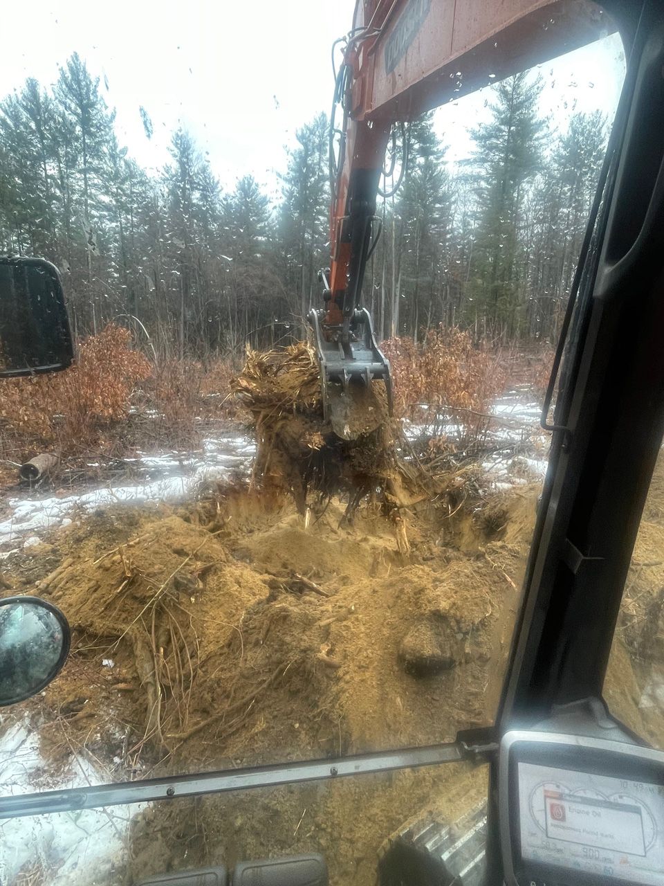 Excavator arm digging in muddy, snowy forest clearing from inside cab view