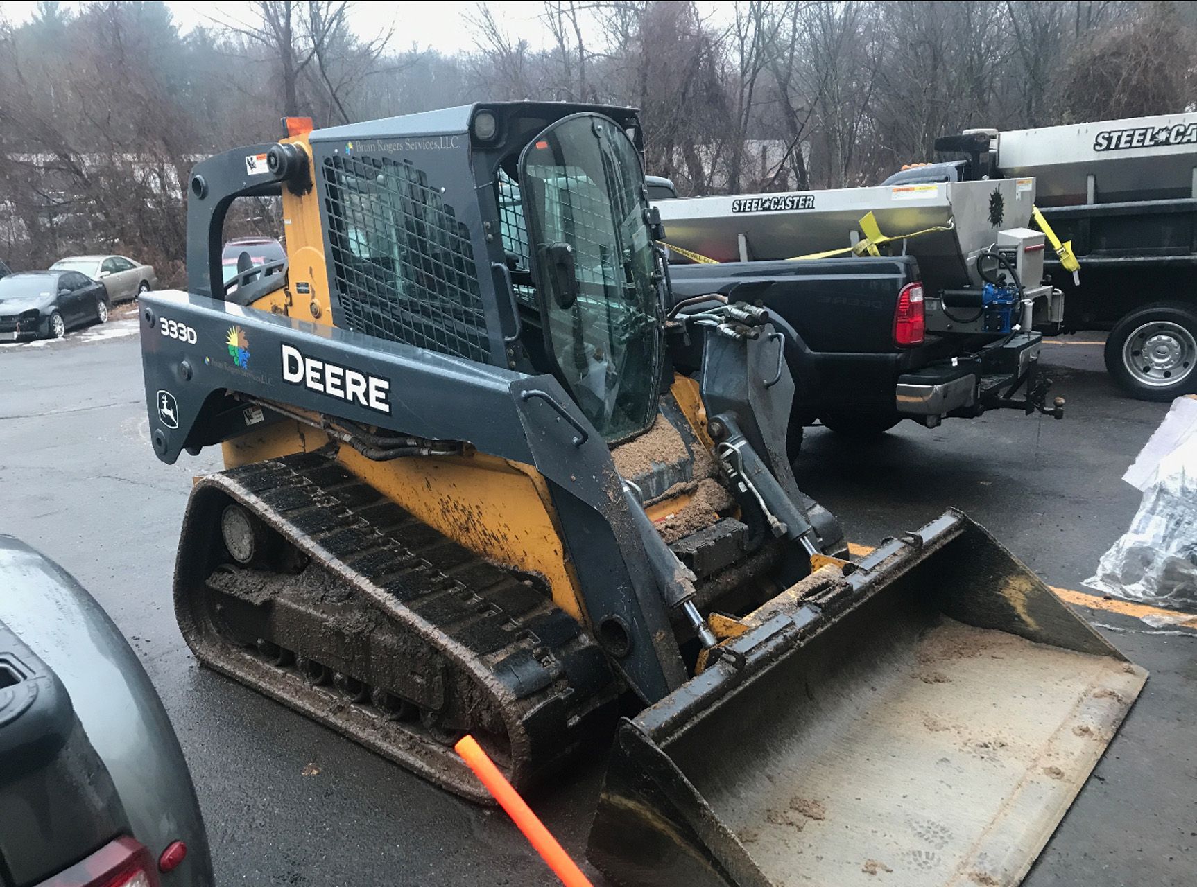 Deere skid steer with front bucket parked in a snowy lot beside trucks and trailers