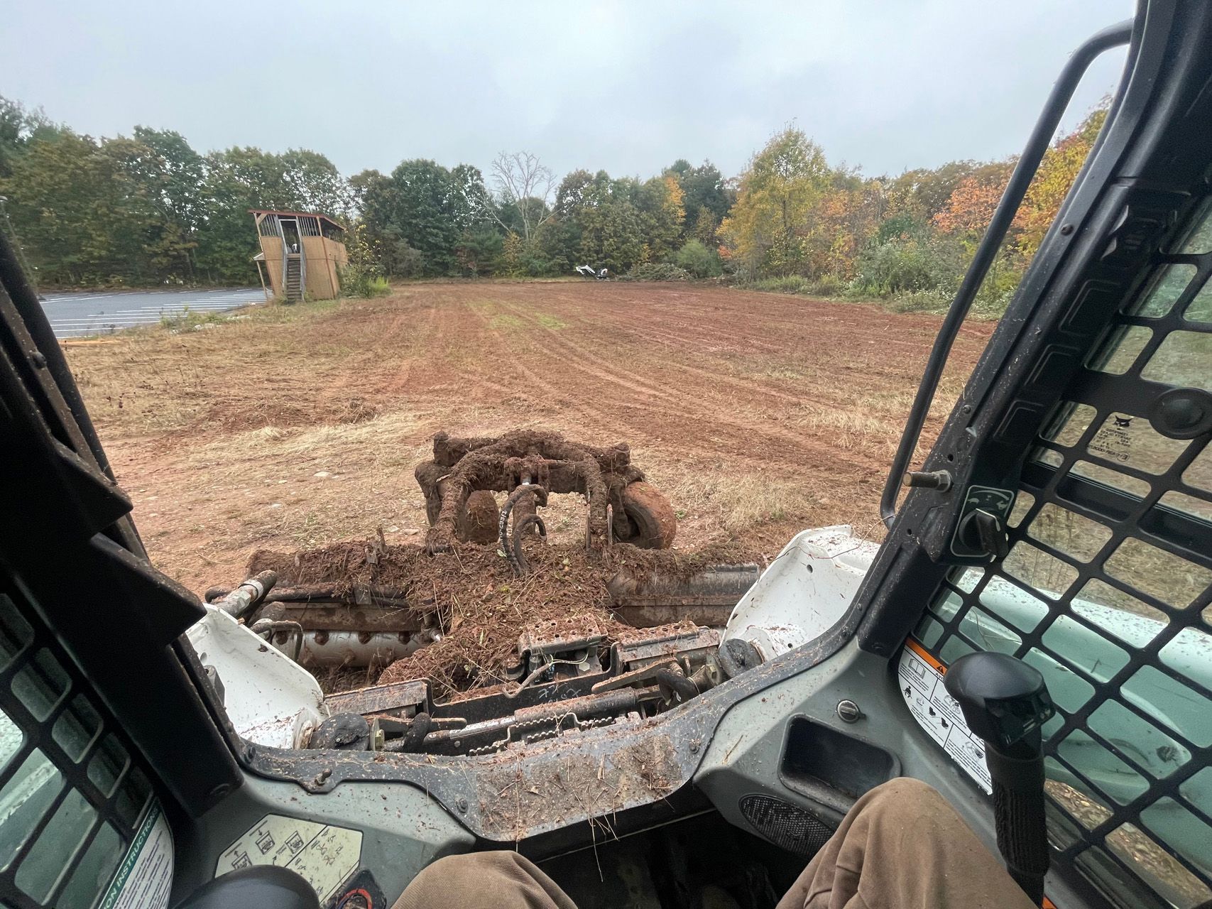 Inside a construction vehicle, a muddy field is being graded with trees in the background.