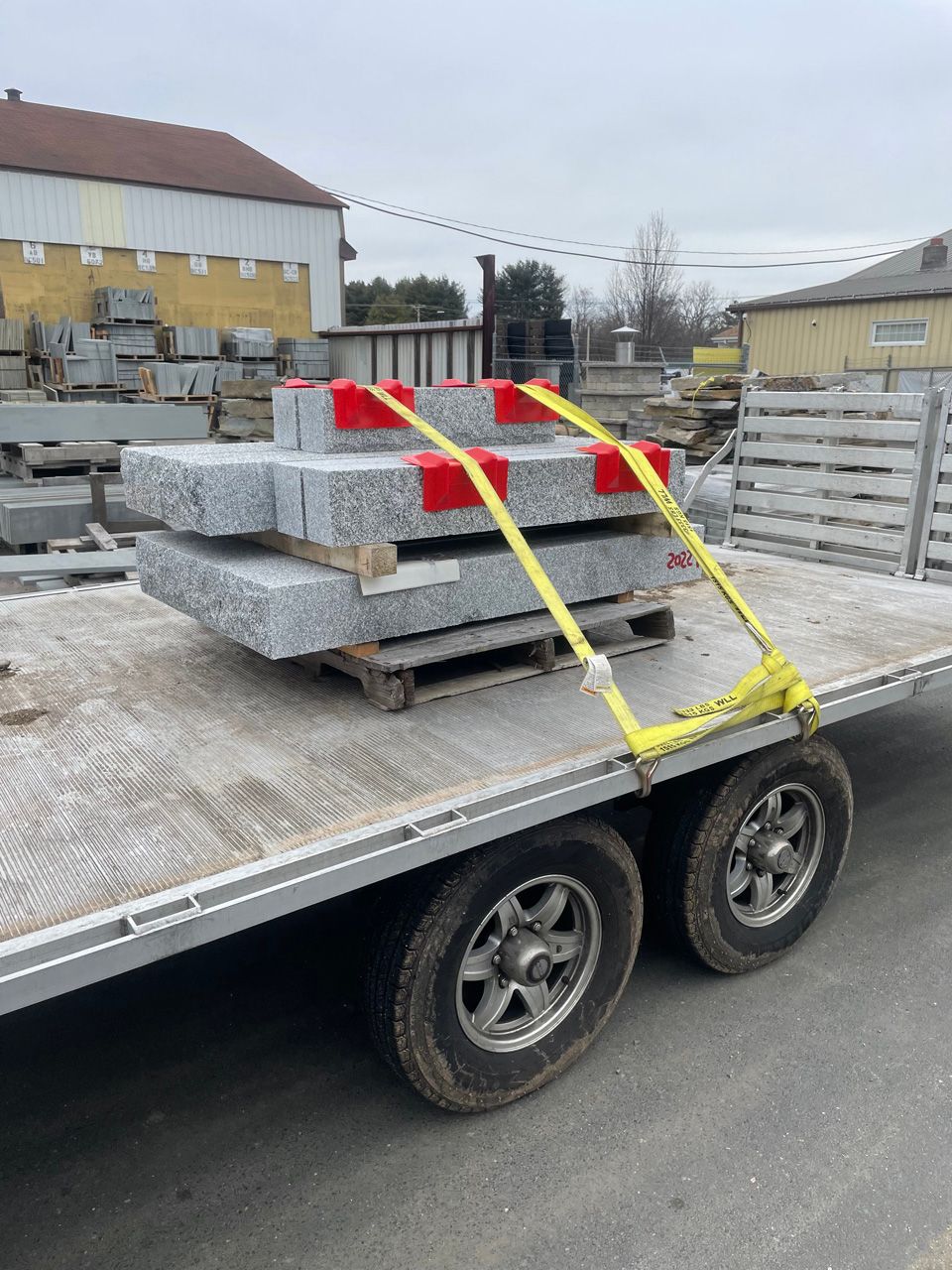 Flatbed trailer loaded with gray stone slabs, secured by yellow straps in a construction yard