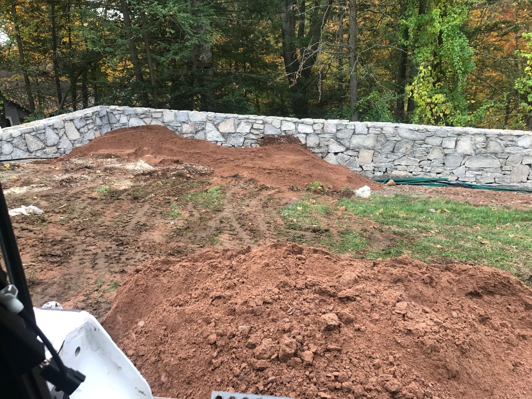 Stone retaining wall beside red dirt piles and patchy grass in a wooded yard