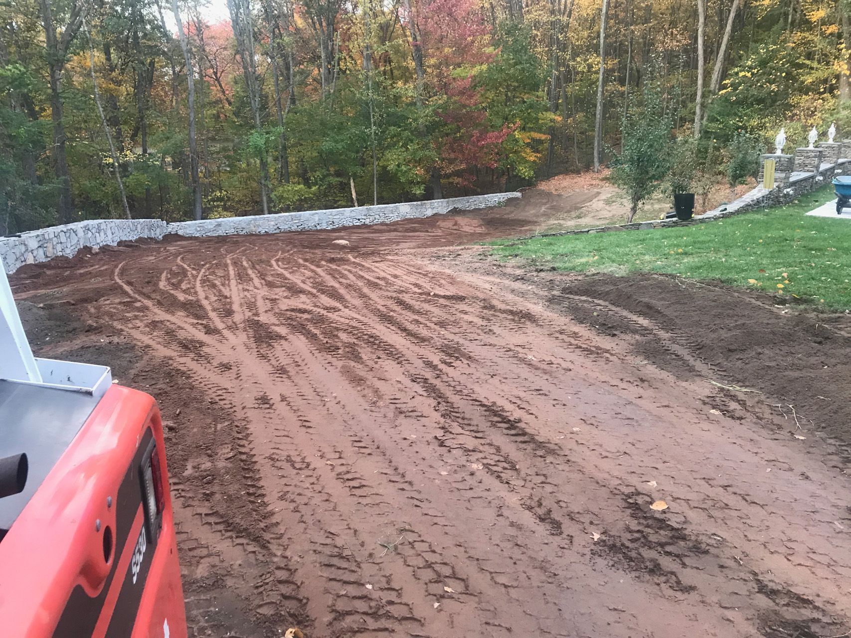 Freshly graded dirt driveway or lot beside a forest, with a red tractor at the left edge.