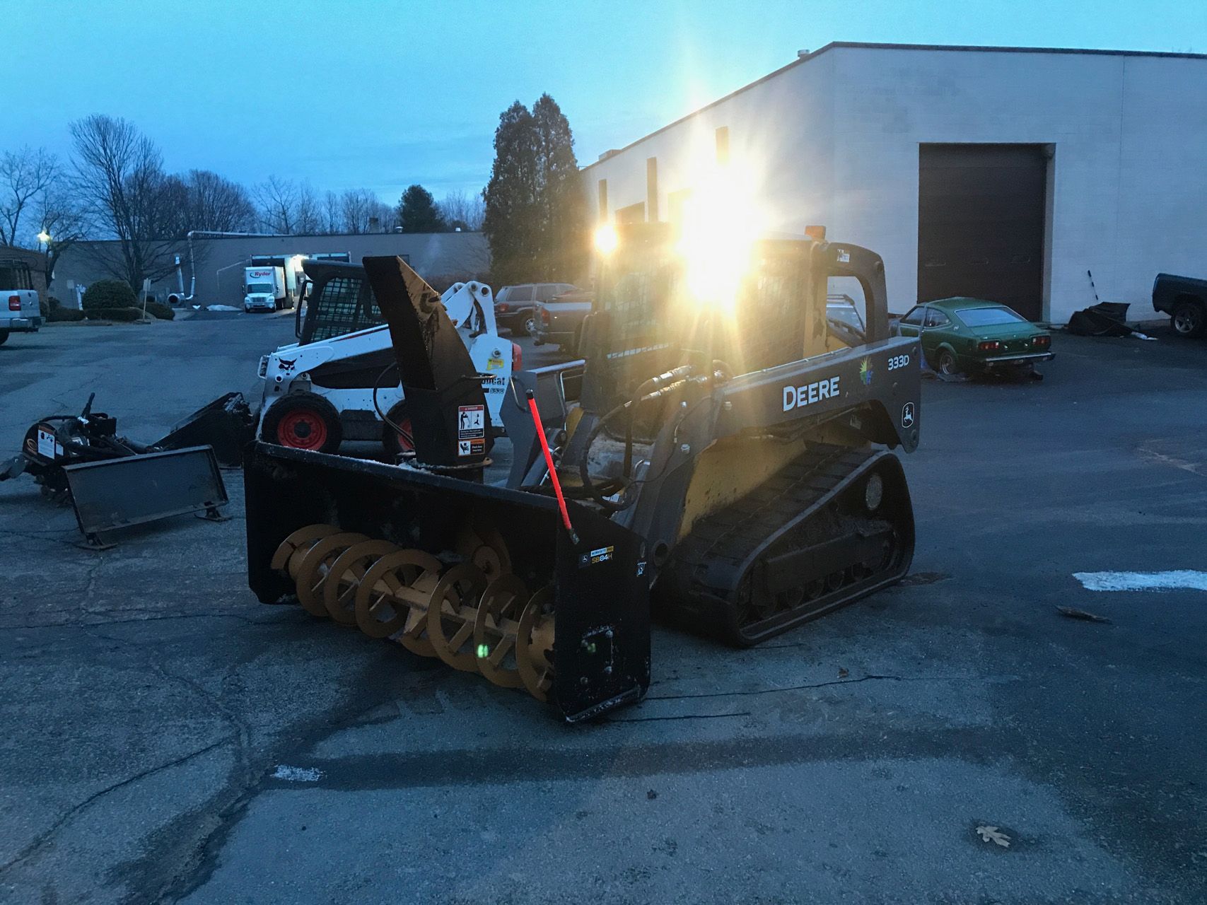 A black tracked skid-steer loader with bright headlights sits in a parking lot at dusk.