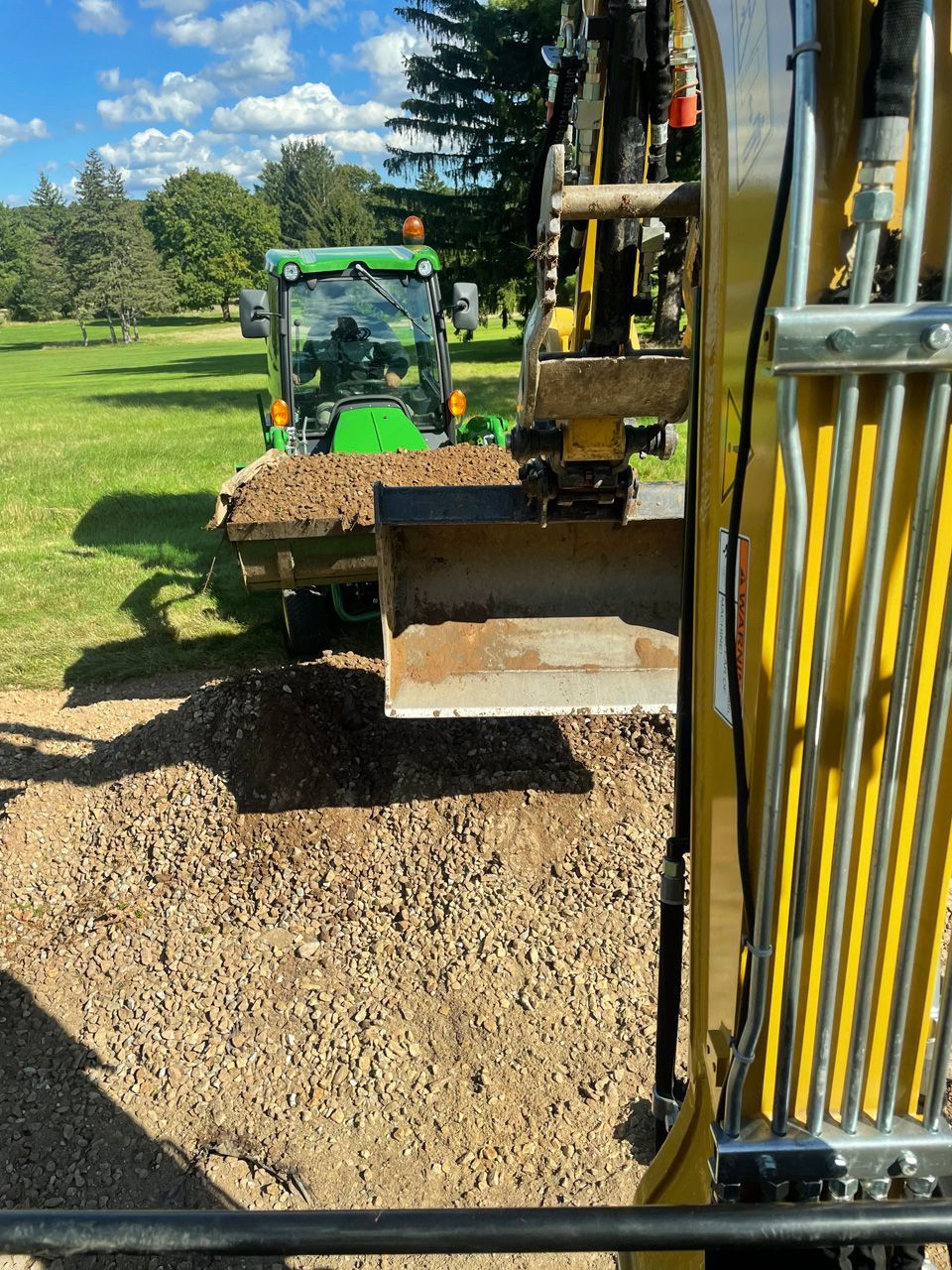 Excavator bucket dumping gravel beside a green tractor in a grassy yard under a blue sky