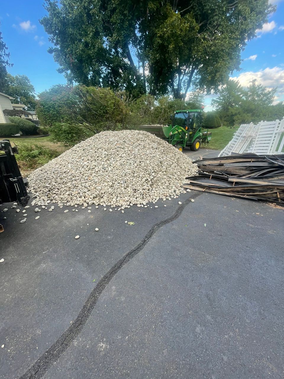 Gravel pile on a driveway beside stacked wood and a green tractor under trees