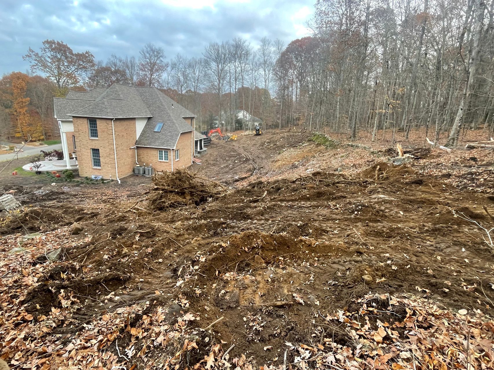 Cleared muddy lot beside a brick house and trees under a cloudy sky