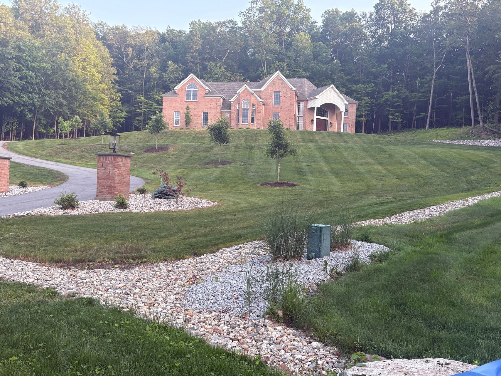 Brick house with arched entry on a grassy hillside, bordered by winding white stone landscaping and trees