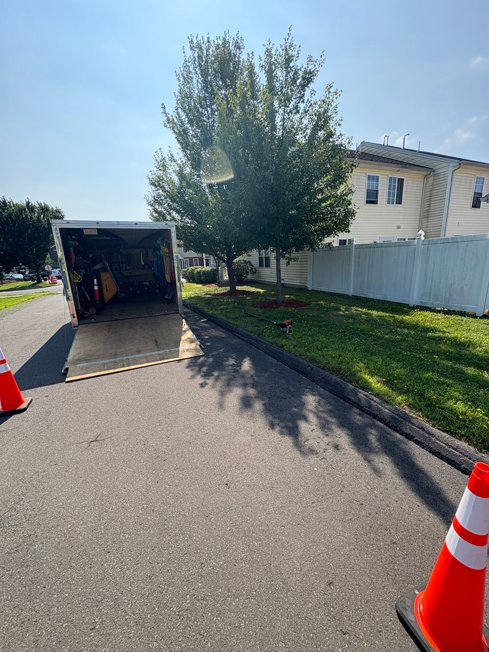 Driveway with open utility trailer, orange cones, and a white house beside a tree-lined street.
