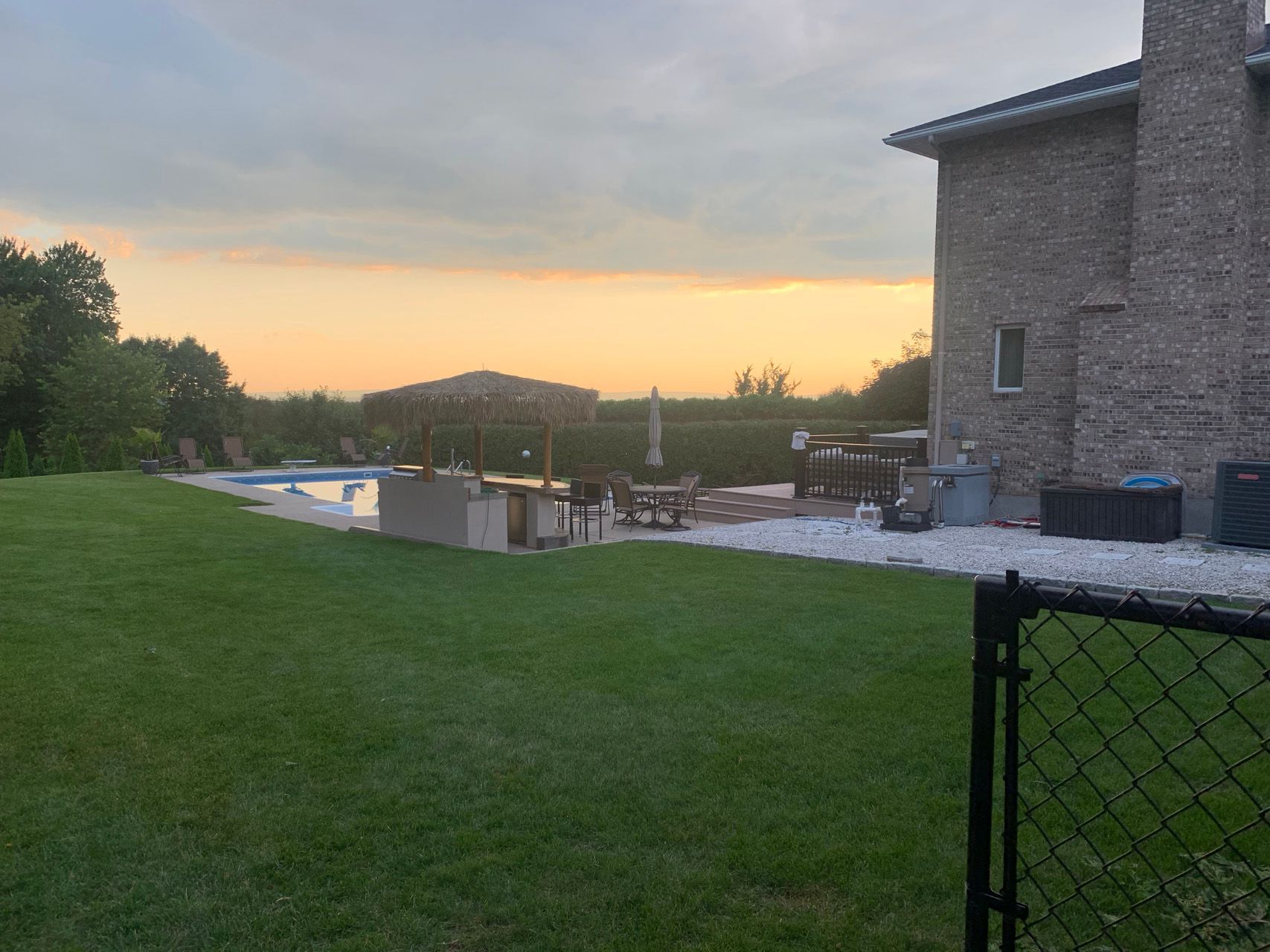 Backyard lawn at sunset beside a stone house, with a pool and patio in the distance