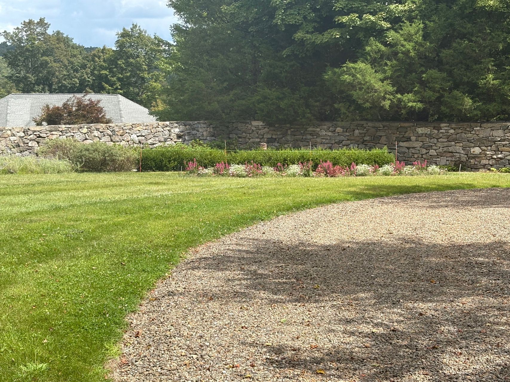 Curved gravel path beside a green lawn, stone wall, and flower bed in a sunny garden.