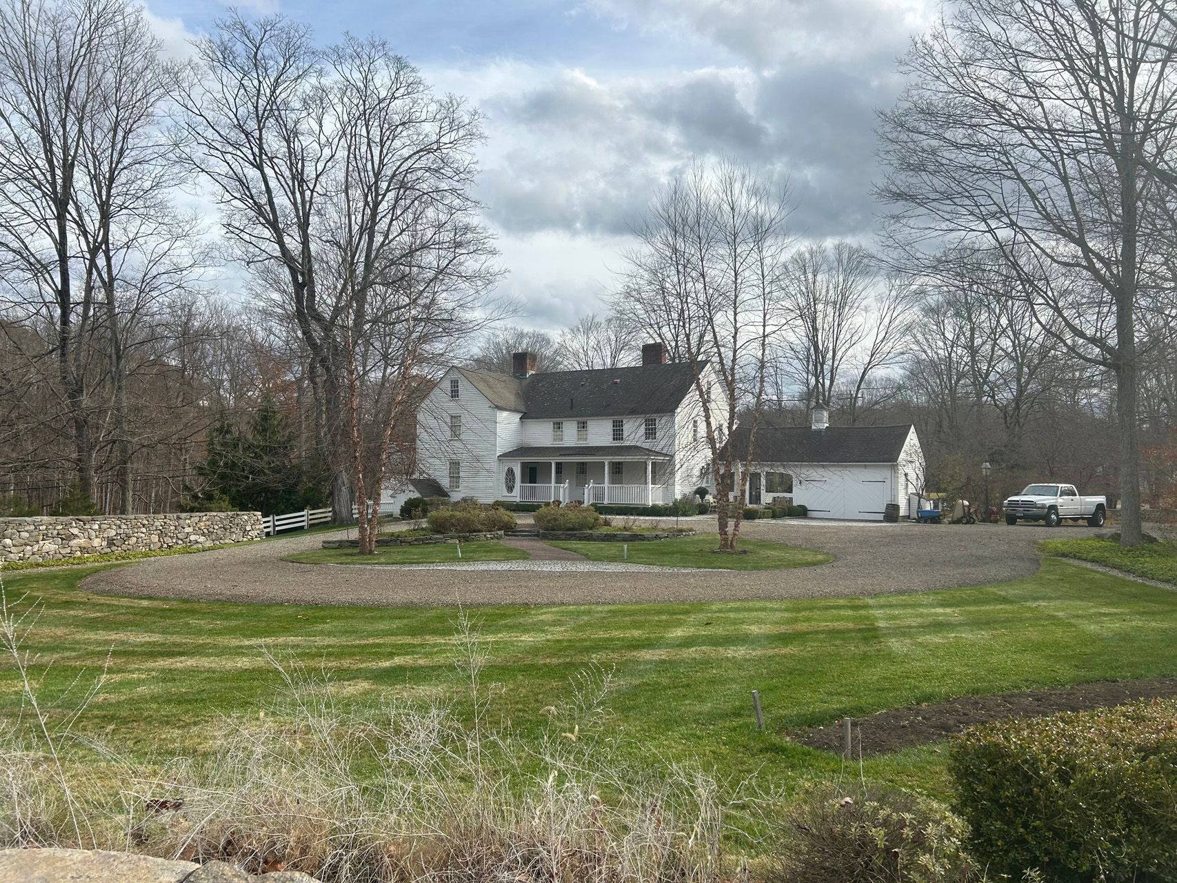 White two-story house with a garage, surrounded by leafless trees and a grassy yard under cloudy skies