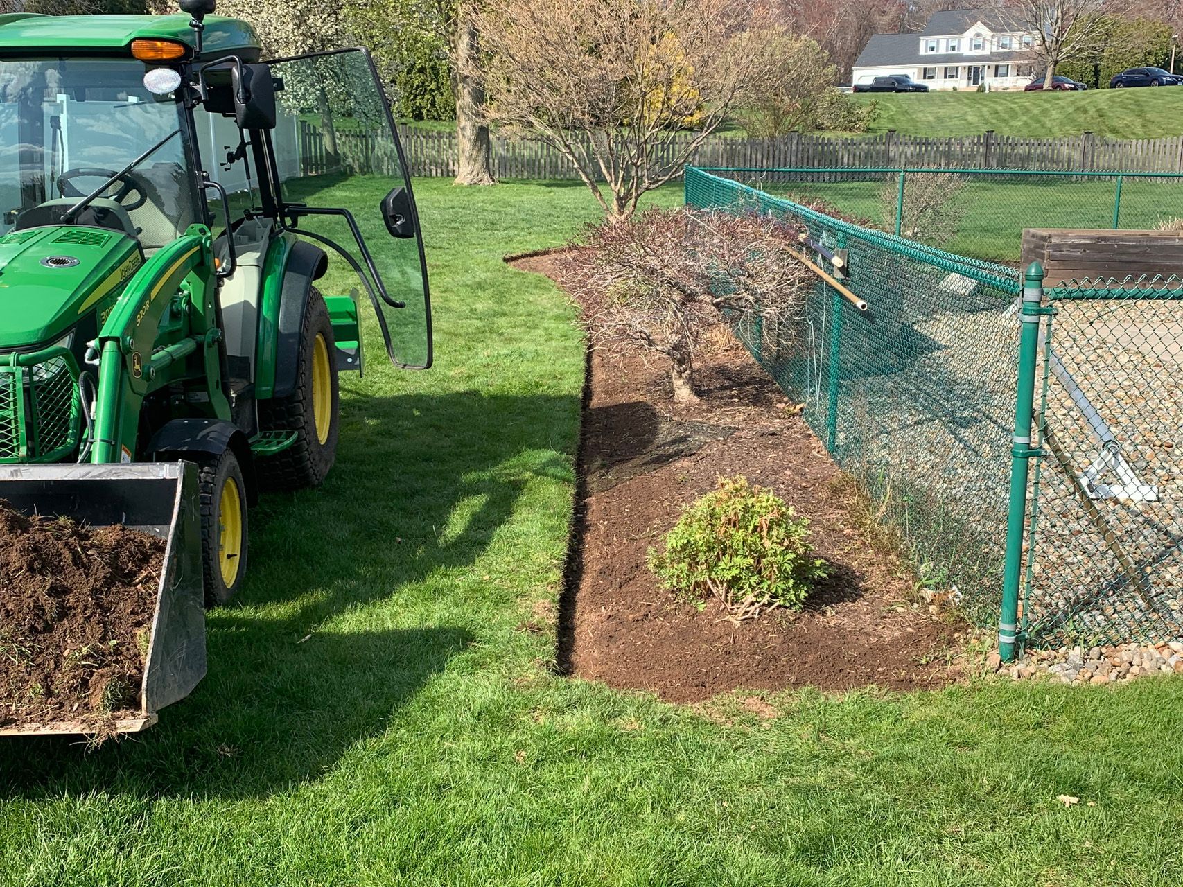 Green tractor with front loader beside a mulched garden bed and chain-link fence in a grassy yard.
