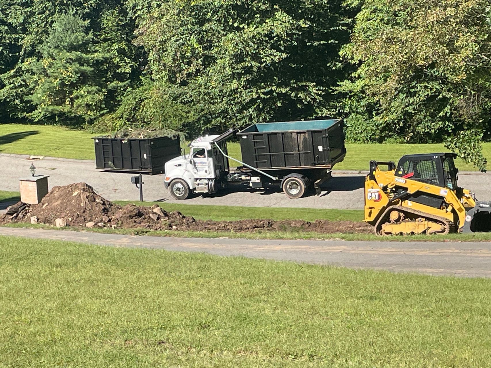 Construction truck, dump trailer, and skid steer near a dirt pile on a grassy roadside.