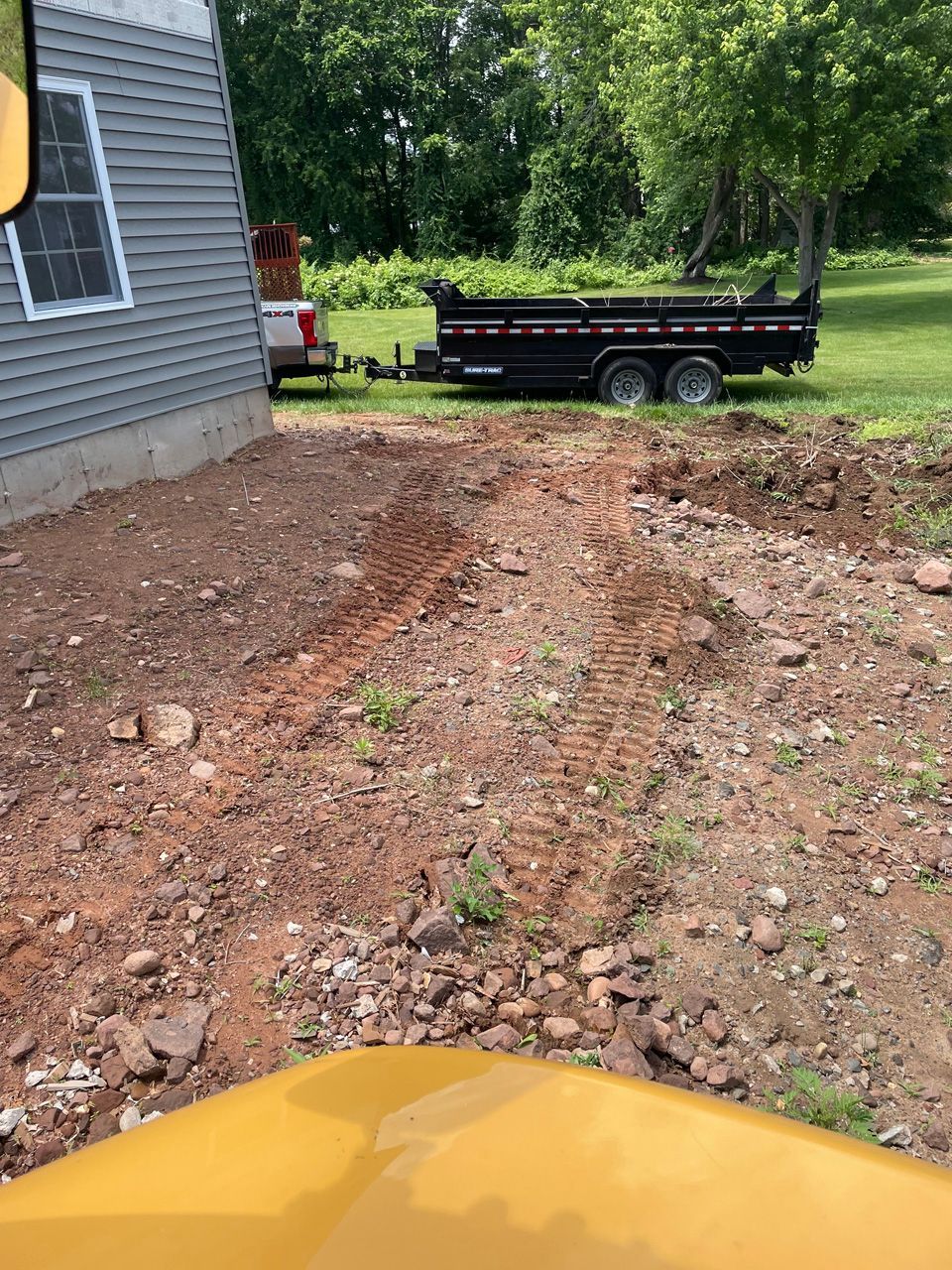 Rough dirt driveway beside a gray house, with a black trailer in the yard and a yellow vehicle hood in foreground