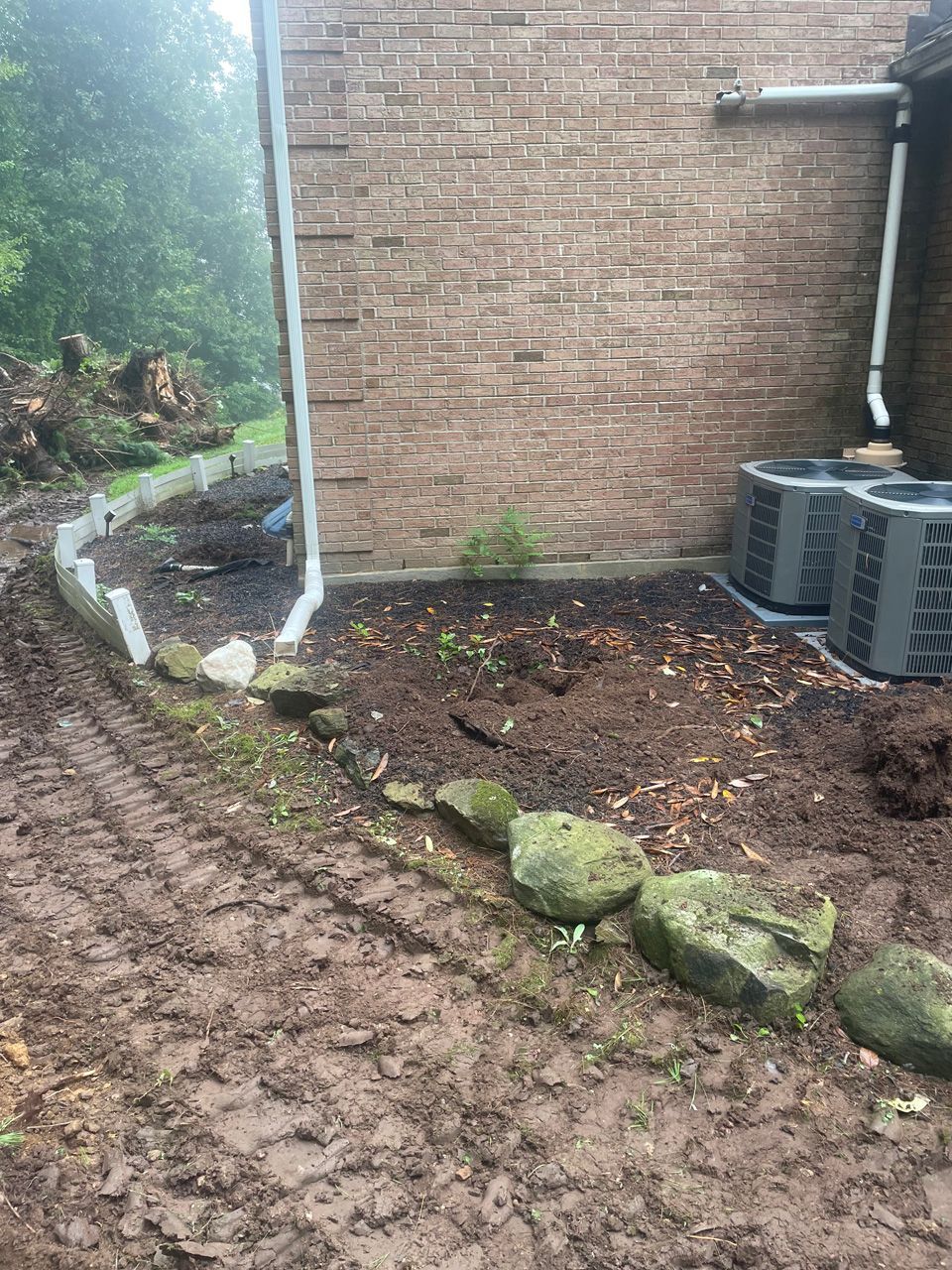 Muddy garden path beside a brick house, with a curved border of rocks and stacked blocks near the wall