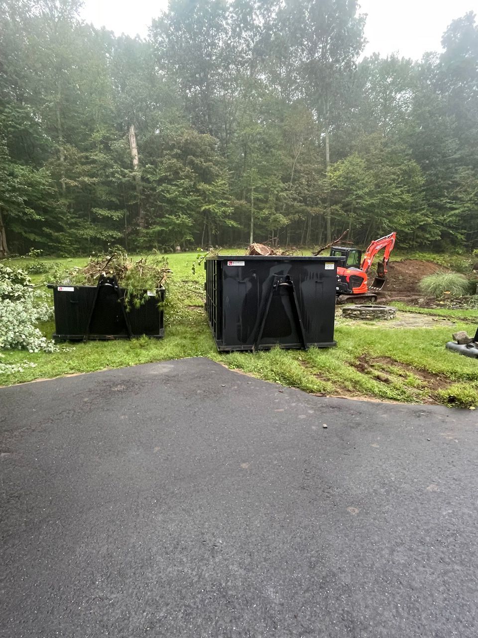 Two black dumpsters on a wet driveway beside a wooded area, with an orange excavator in the background.