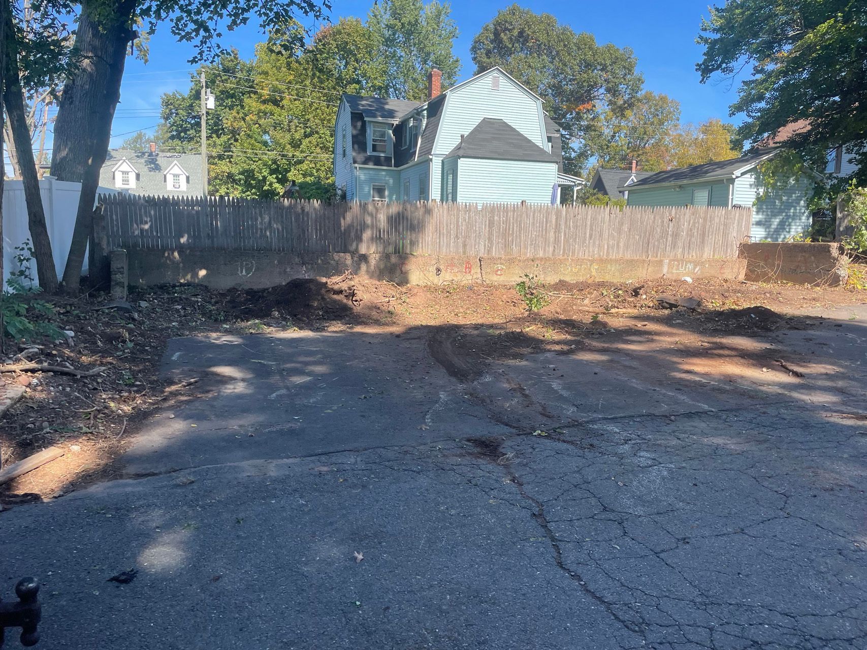 Vacant lot with cracked asphalt and dirt, bordered by a wooden fence and houses under trees.