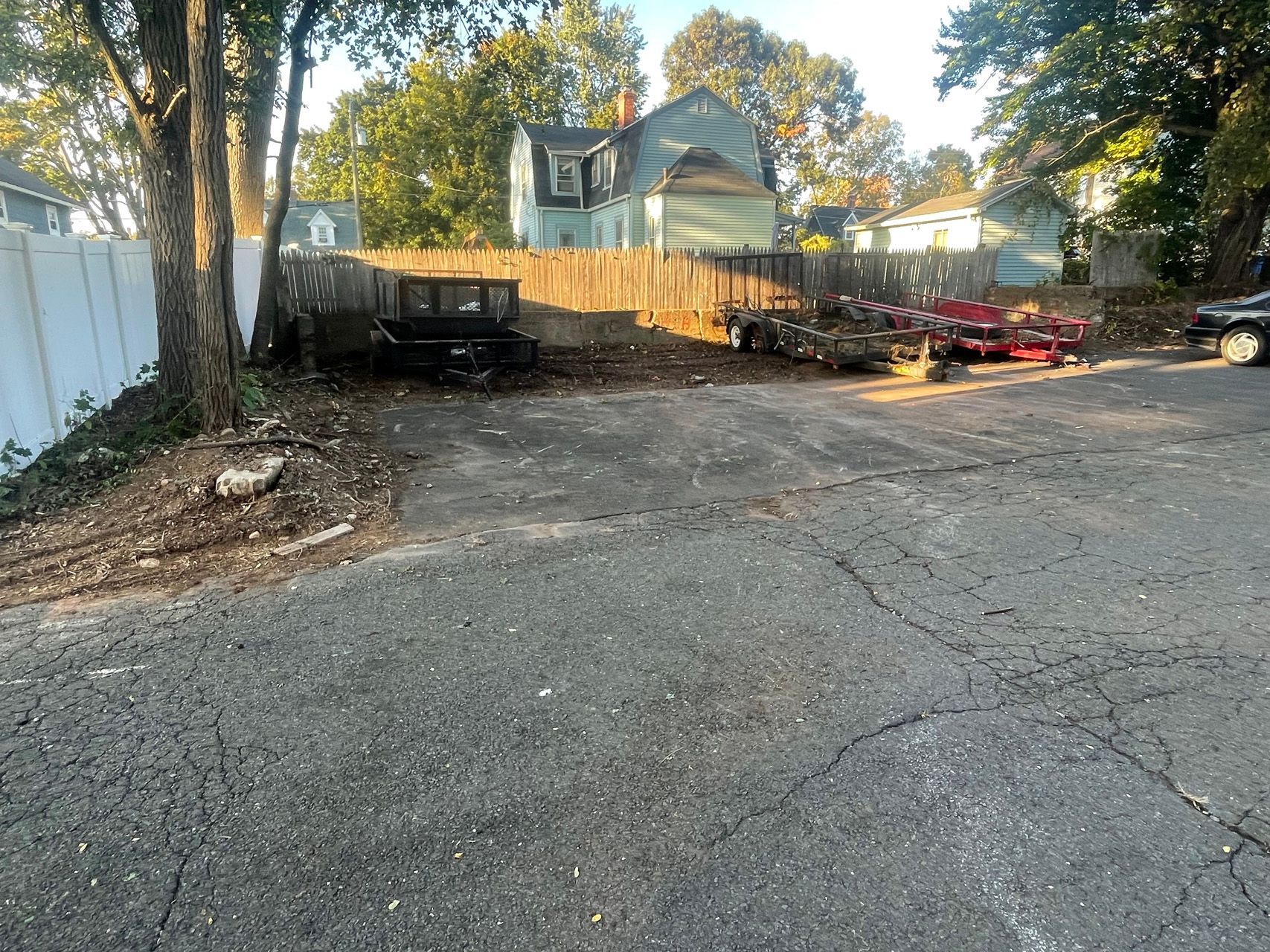 Gravel lot with parked trailers, piles of debris, and trees beside a wooden fence and houses