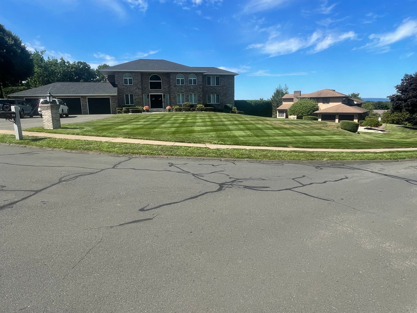 Suburban street with large brick house, manicured lawn, driveway, and blue sky on a sunny day