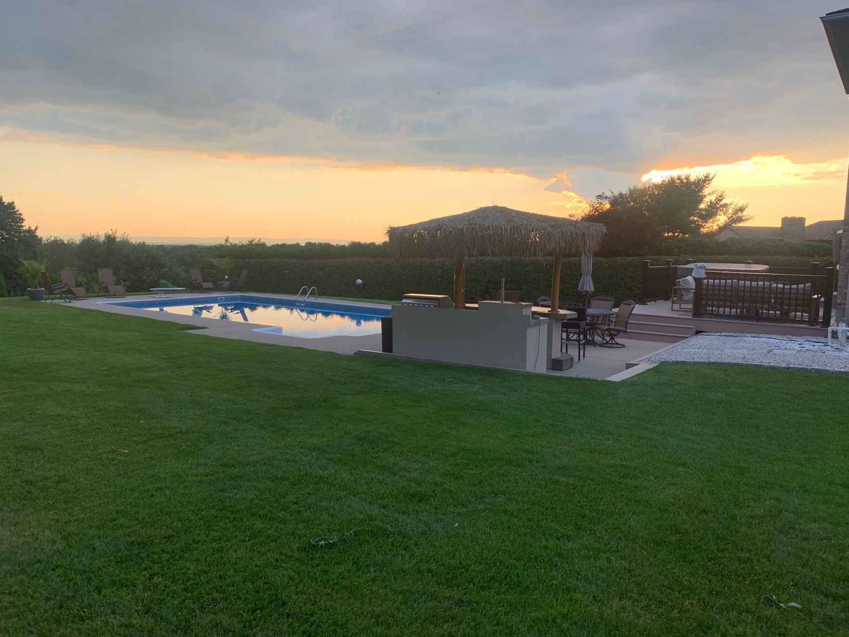 Sunset view of a backyard with green lawn, swimming pool, and patio seating under a covered pergola