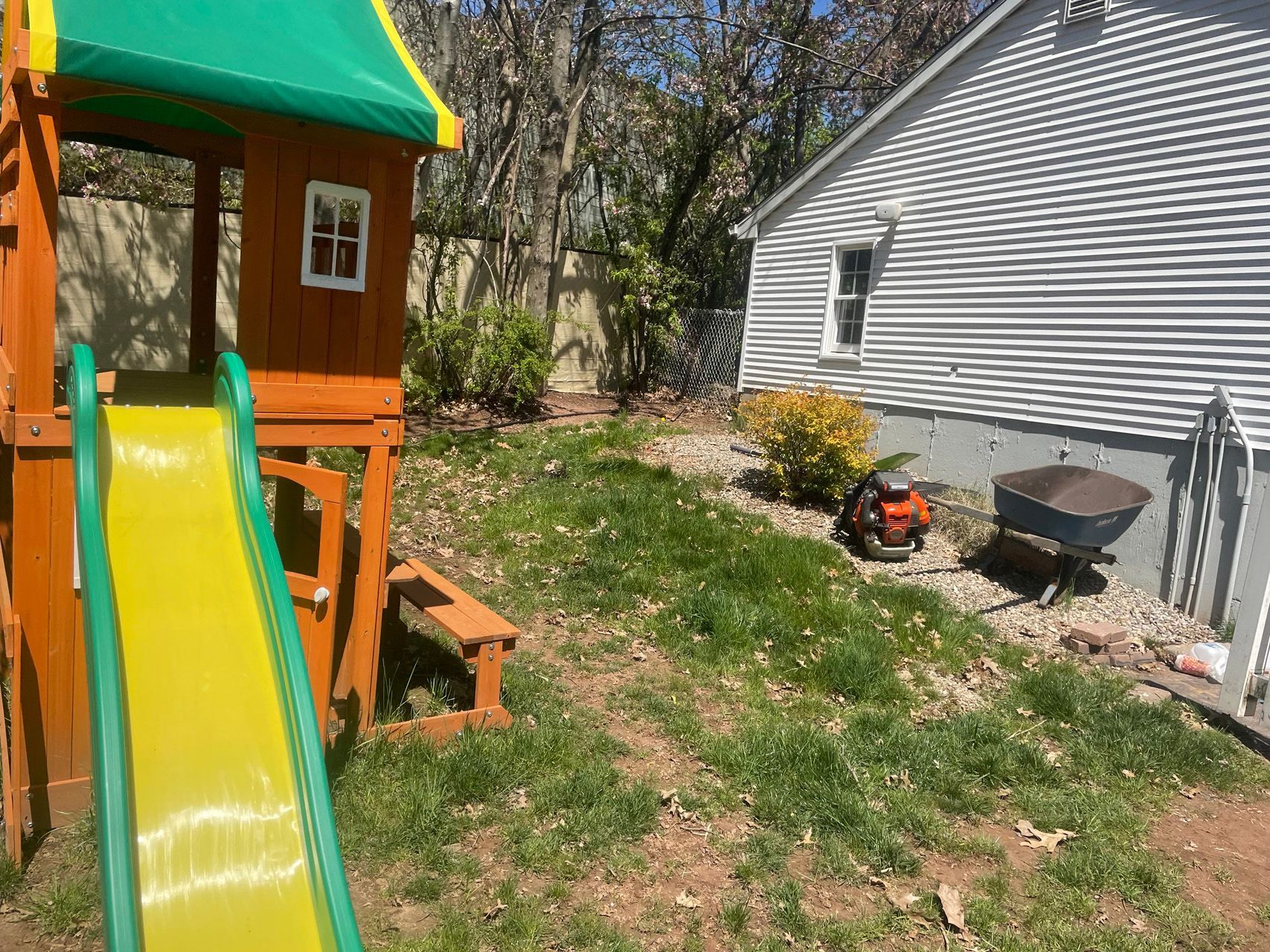 Backyard playset with yellow slide beside a gray house, trees, and a small grill on grass.