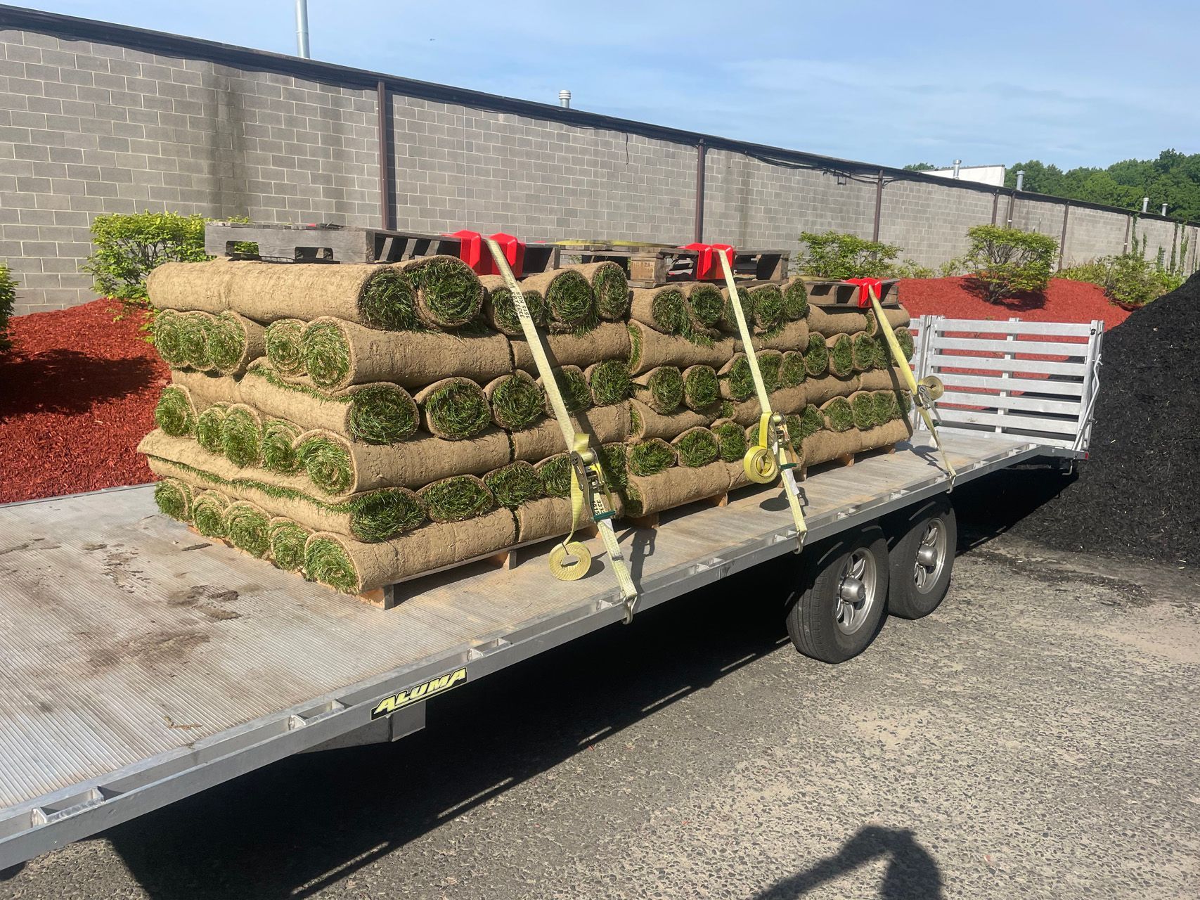 Utility trailer loaded with stacked sod rolls secured by straps in a sunny parking lot
