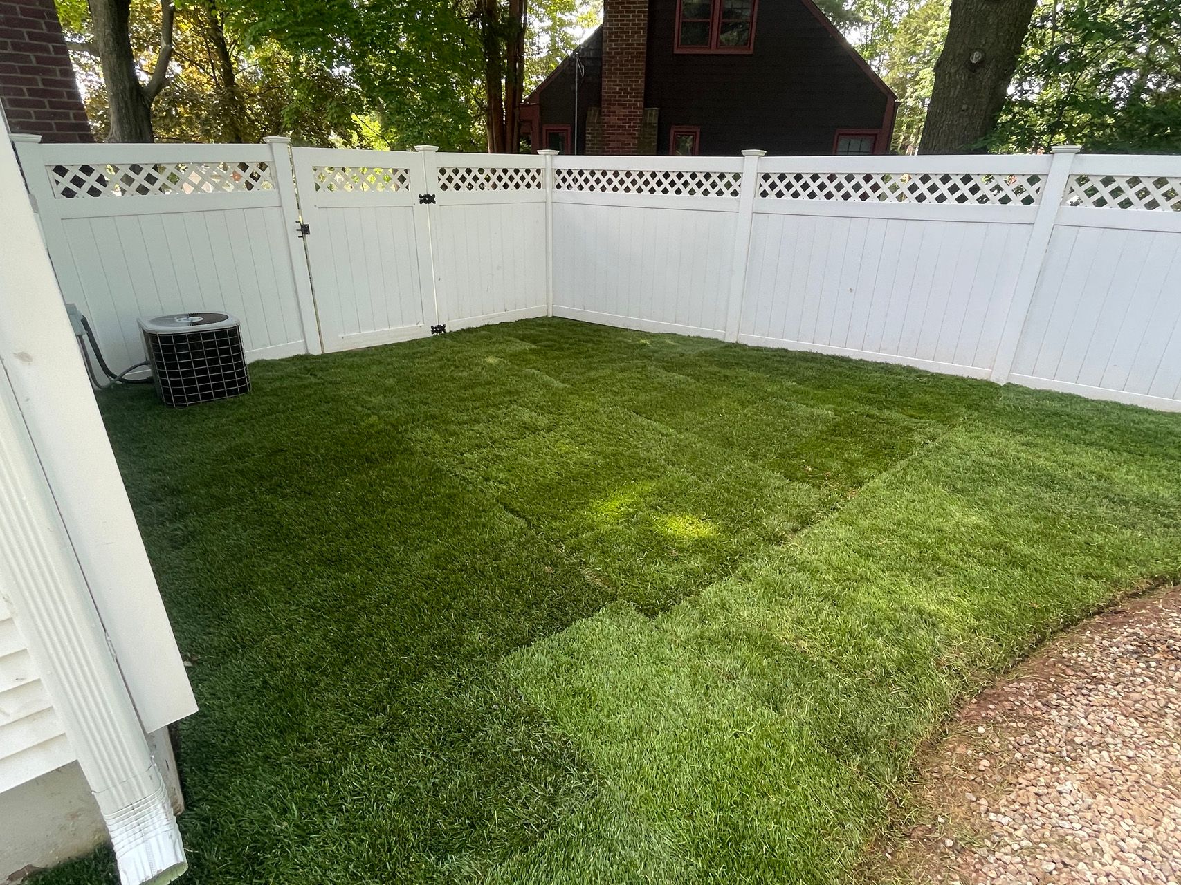 Small fenced backyard with green lawn, white privacy fence, trees, and a dark storage bin in the corner