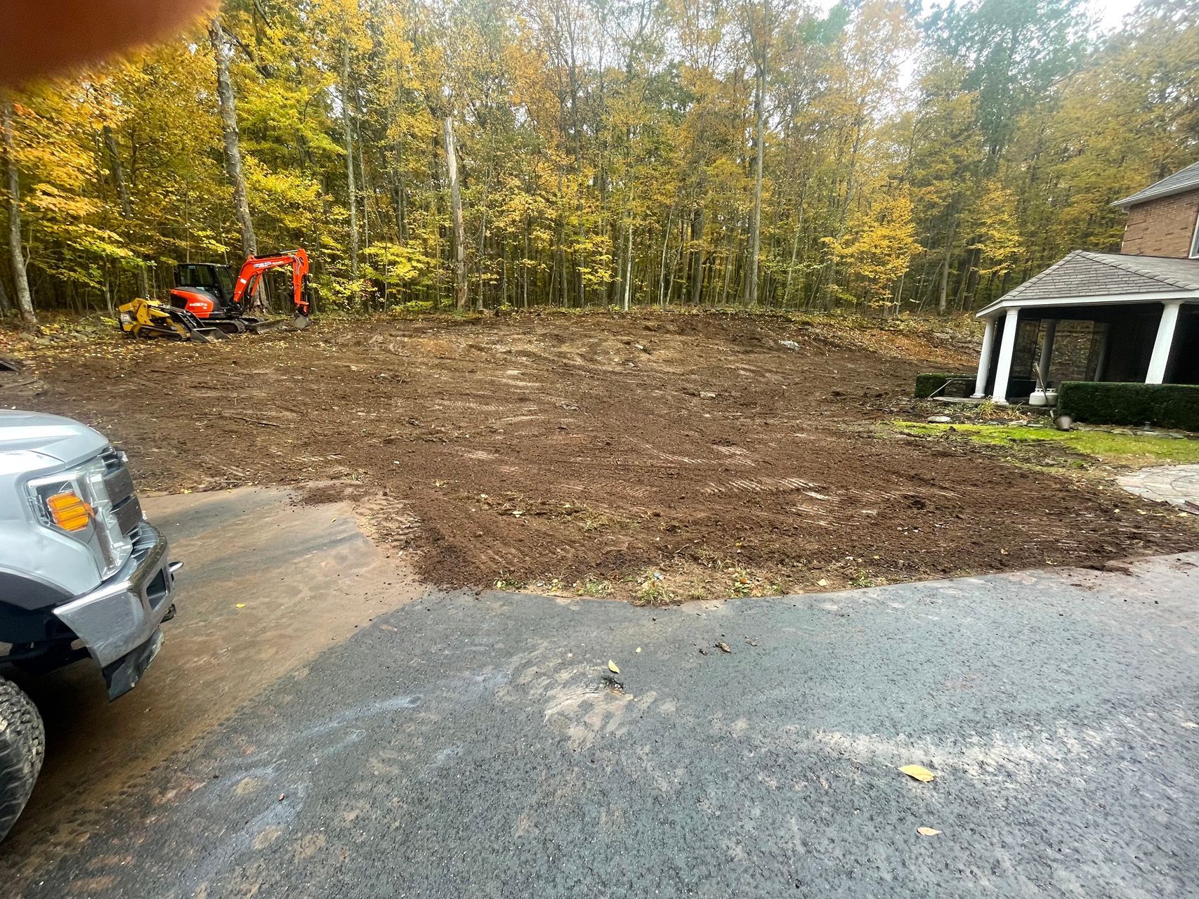 Cleared dirt lot beside a driveway, with an orange excavator, trees, and a small white gazebo