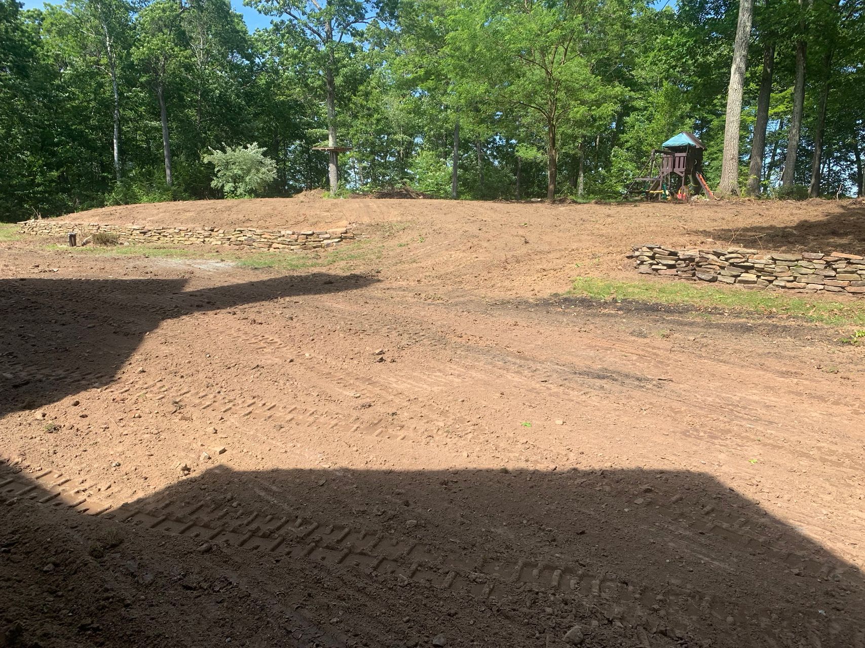 Sunlit dirt clearing with trees along the edge and a small play structure in the distance