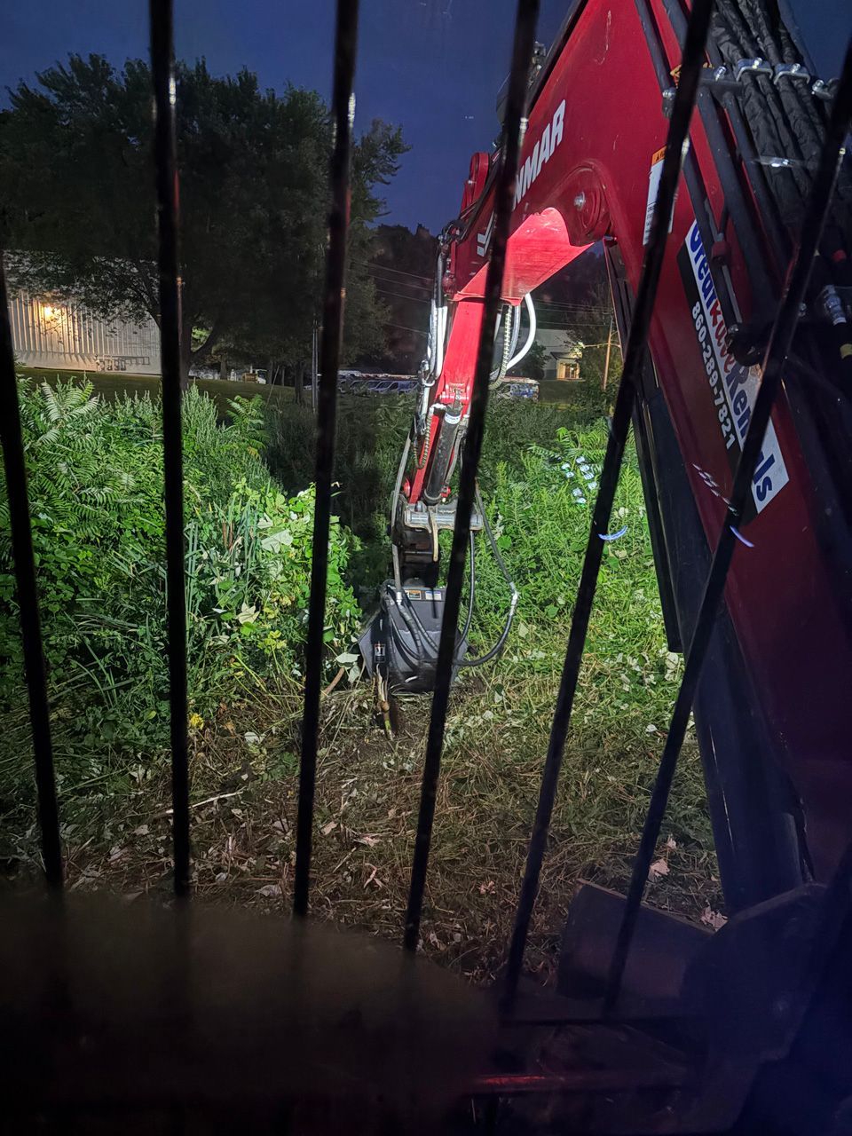 Red excavator working beside a dark fence at night, clearing dense green brush near a lit building