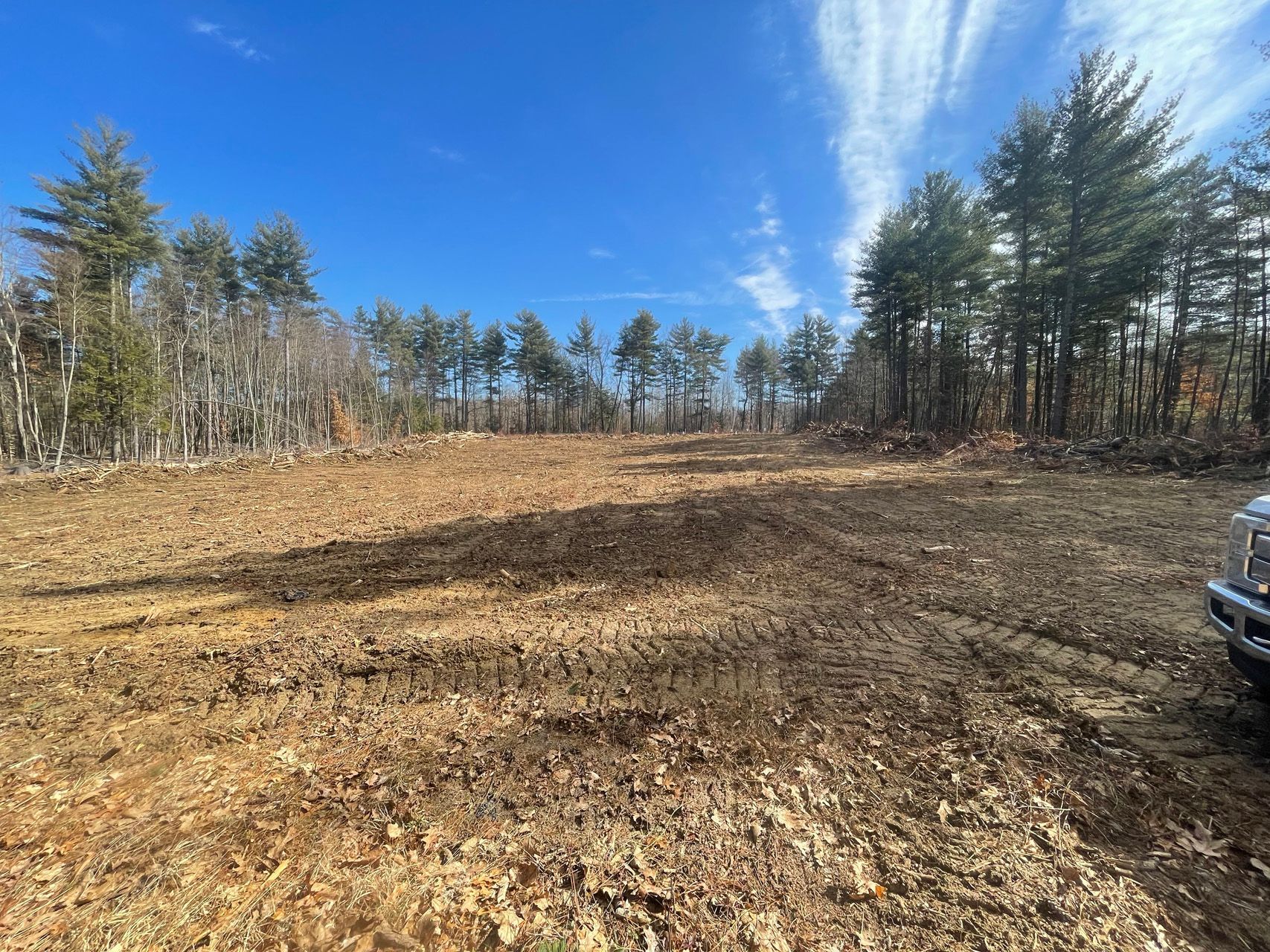 Cleared dirt lot bordered by trees under a bright blue sky, with part of a vehicle at right.