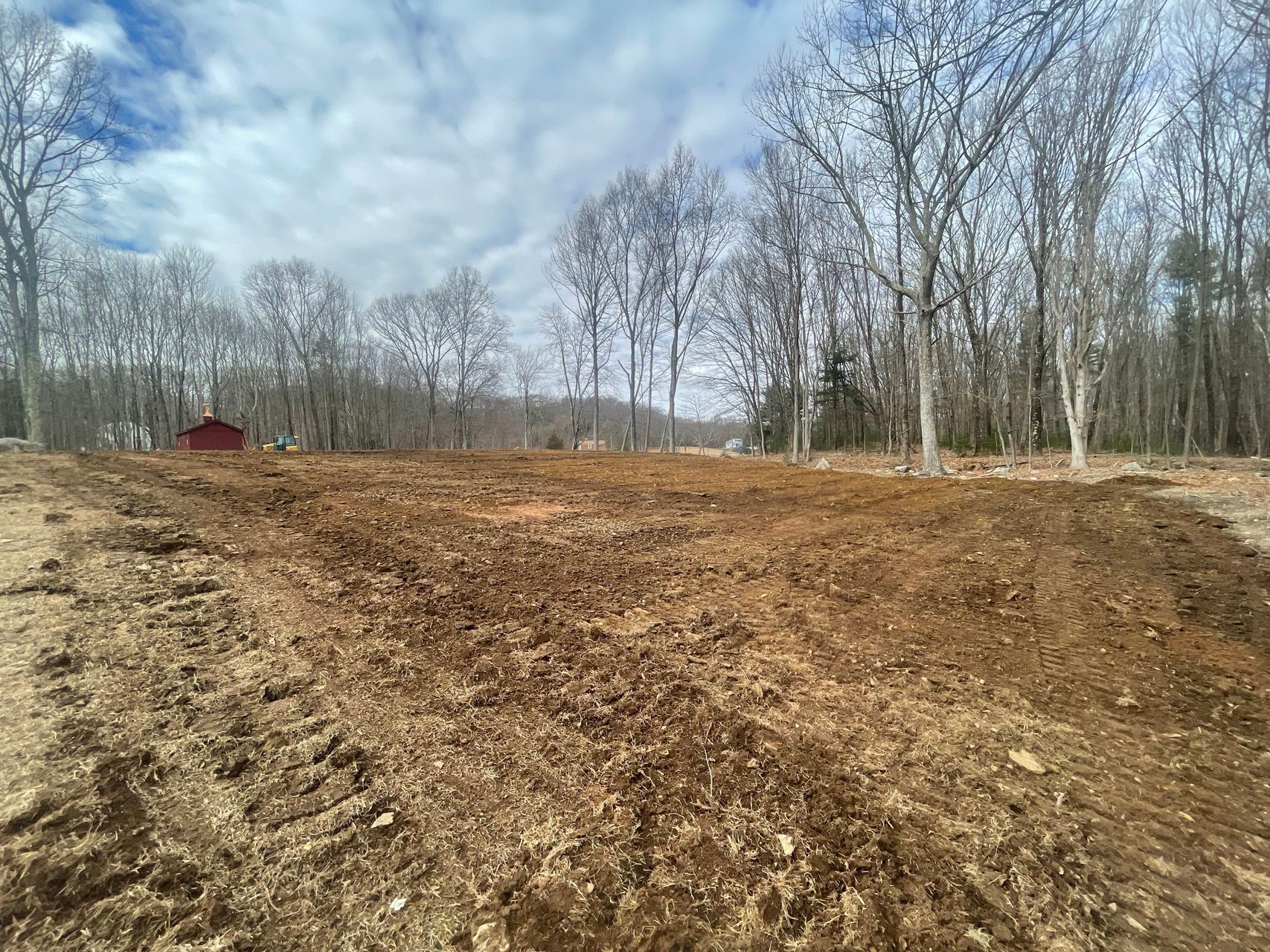 Bare dirt field beside leafless trees under a cloudy sky, with a small red shed in the distance