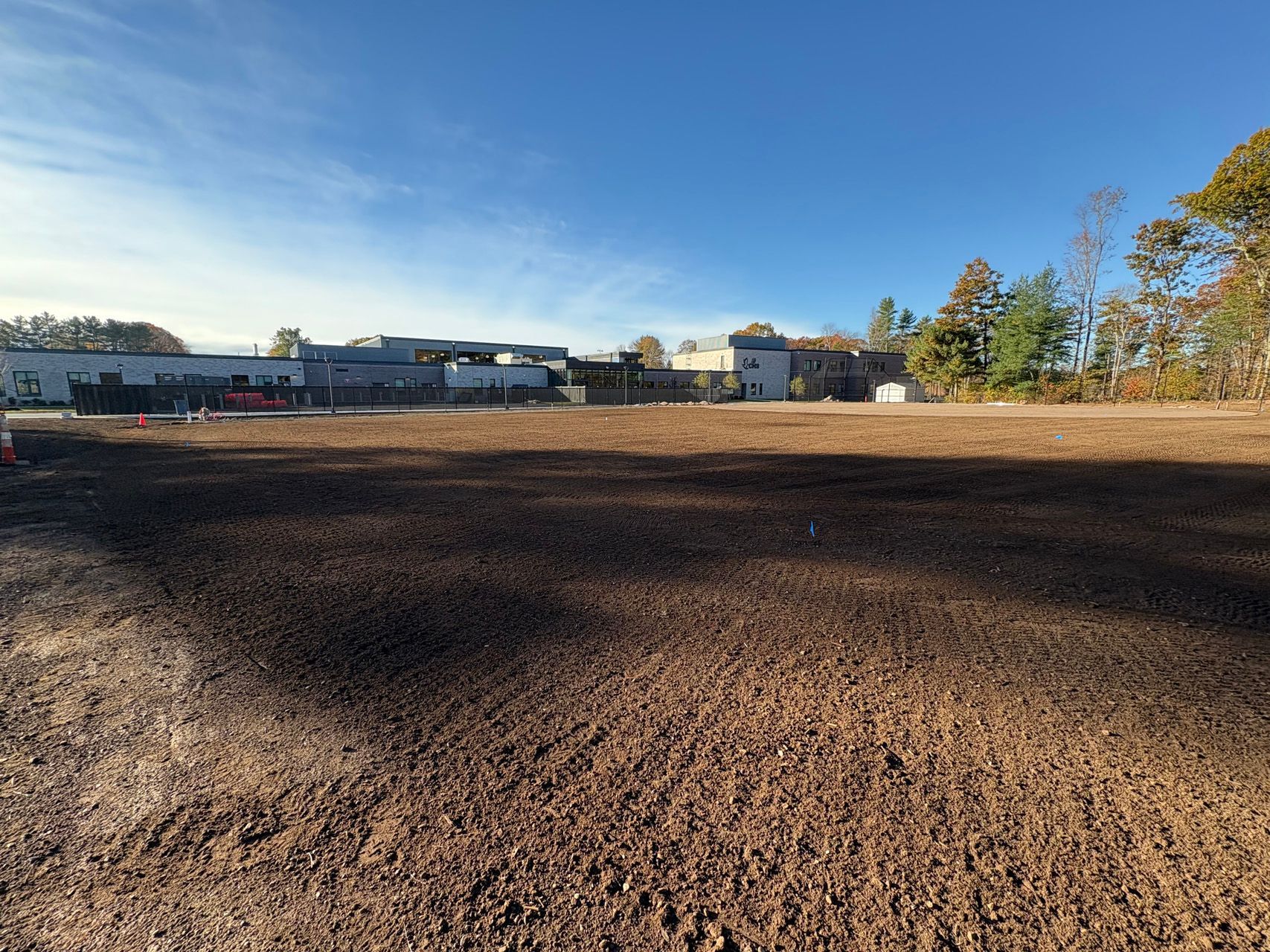 Wide dirt field under a clear blue sky, with buildings and autumn trees in the distance.