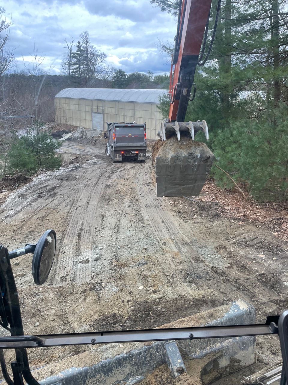 Construction truck and excavator on a muddy dirt road near a building and trees.