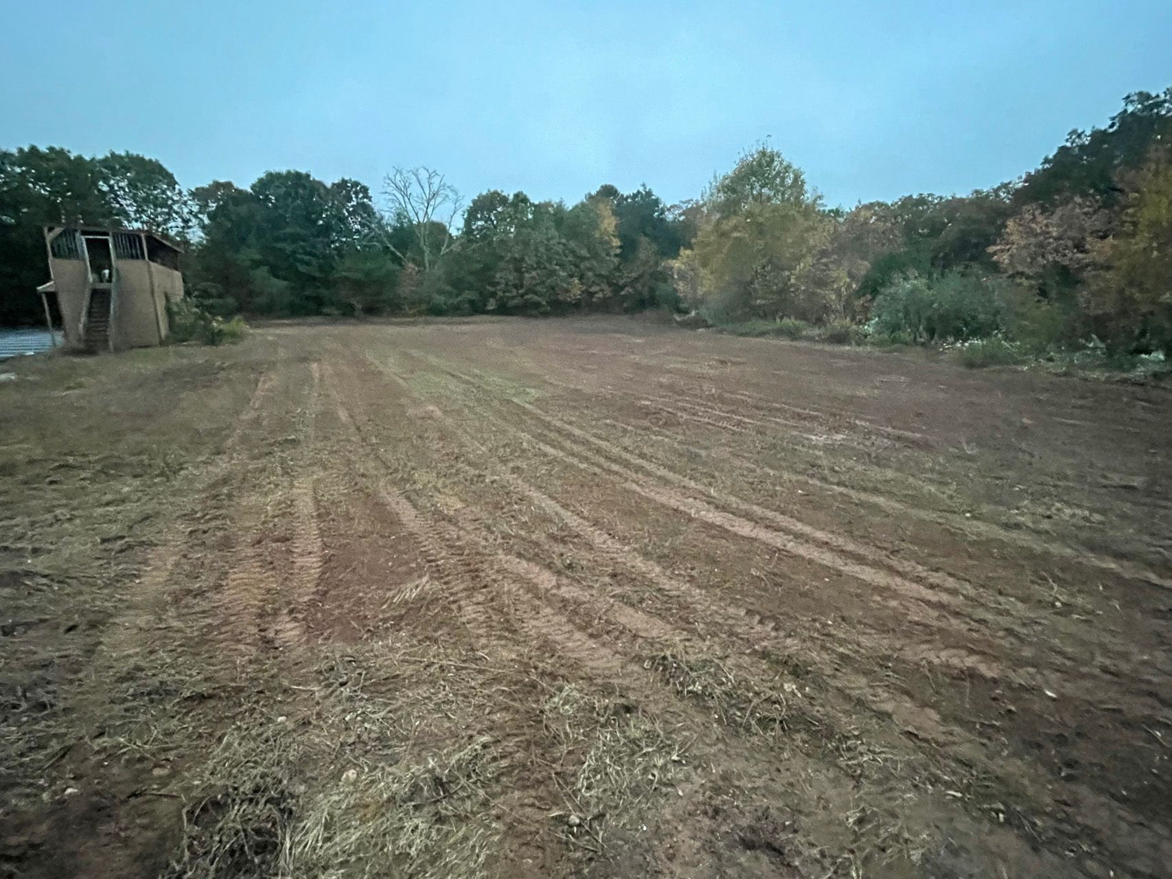 Plowed dirt field bordered by trees and a small building under a cloudy sky