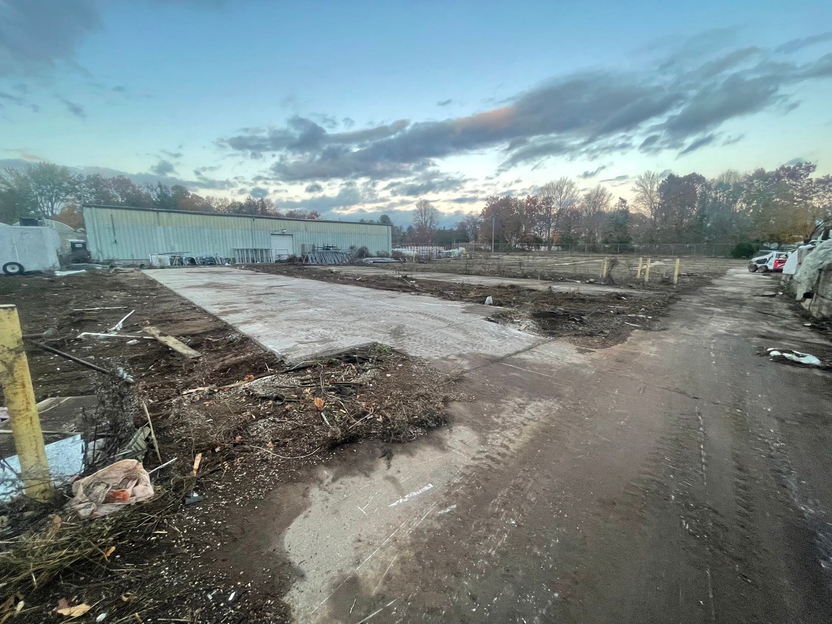 Dirt road and muddy lot beside a long white industrial building at dusk under a cloudy sky