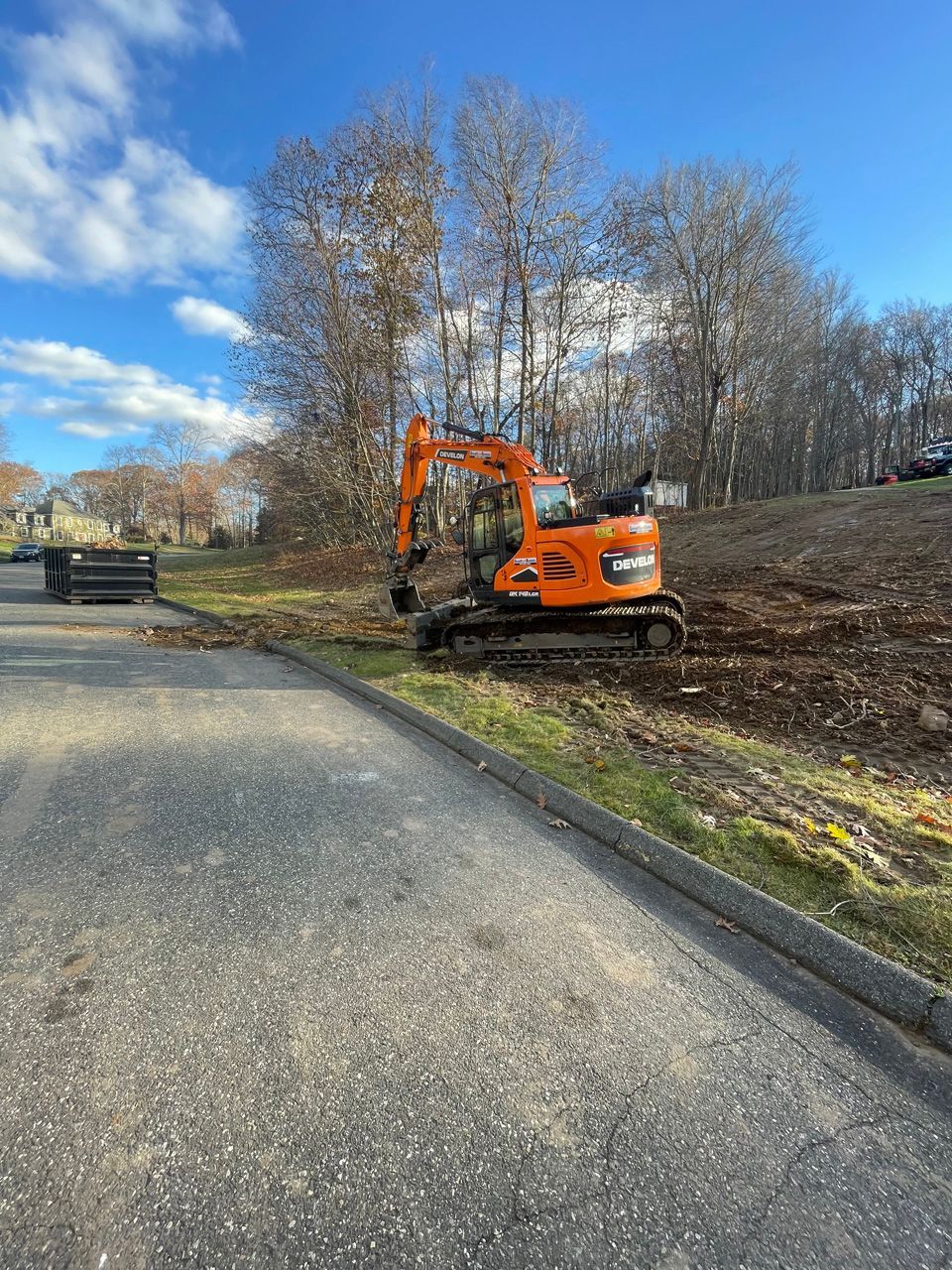 Orange excavator beside a road under a blue sky, clearing dirt on a slope.