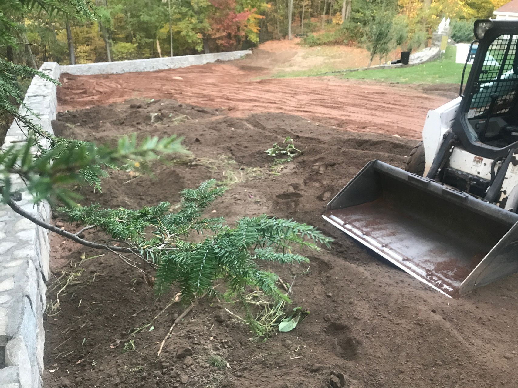 Construction site with skid steer bucket smoothing brown soil beside a stone wall and trees