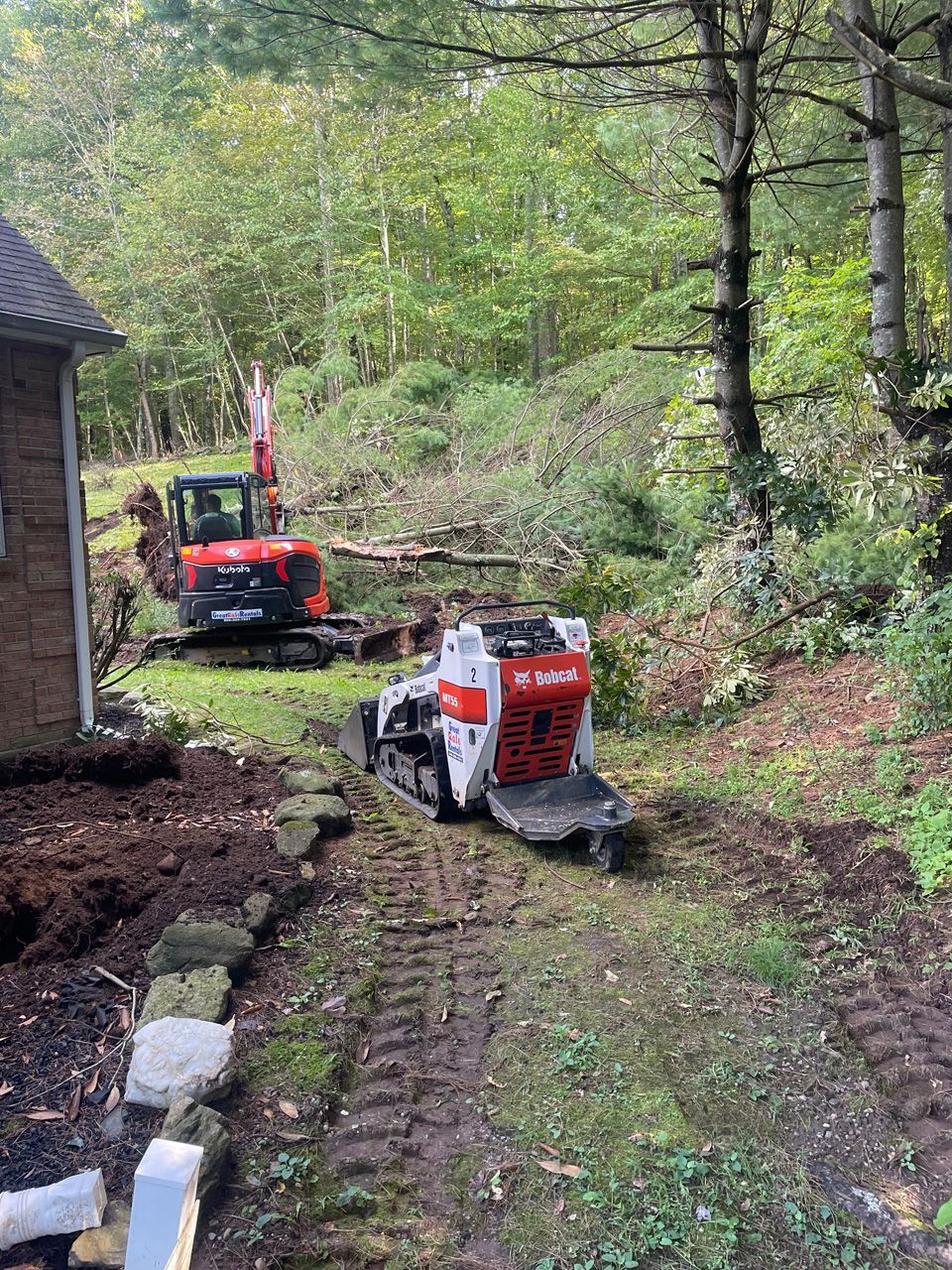 Two small excavators on a muddy path beside a house in a wooded yard