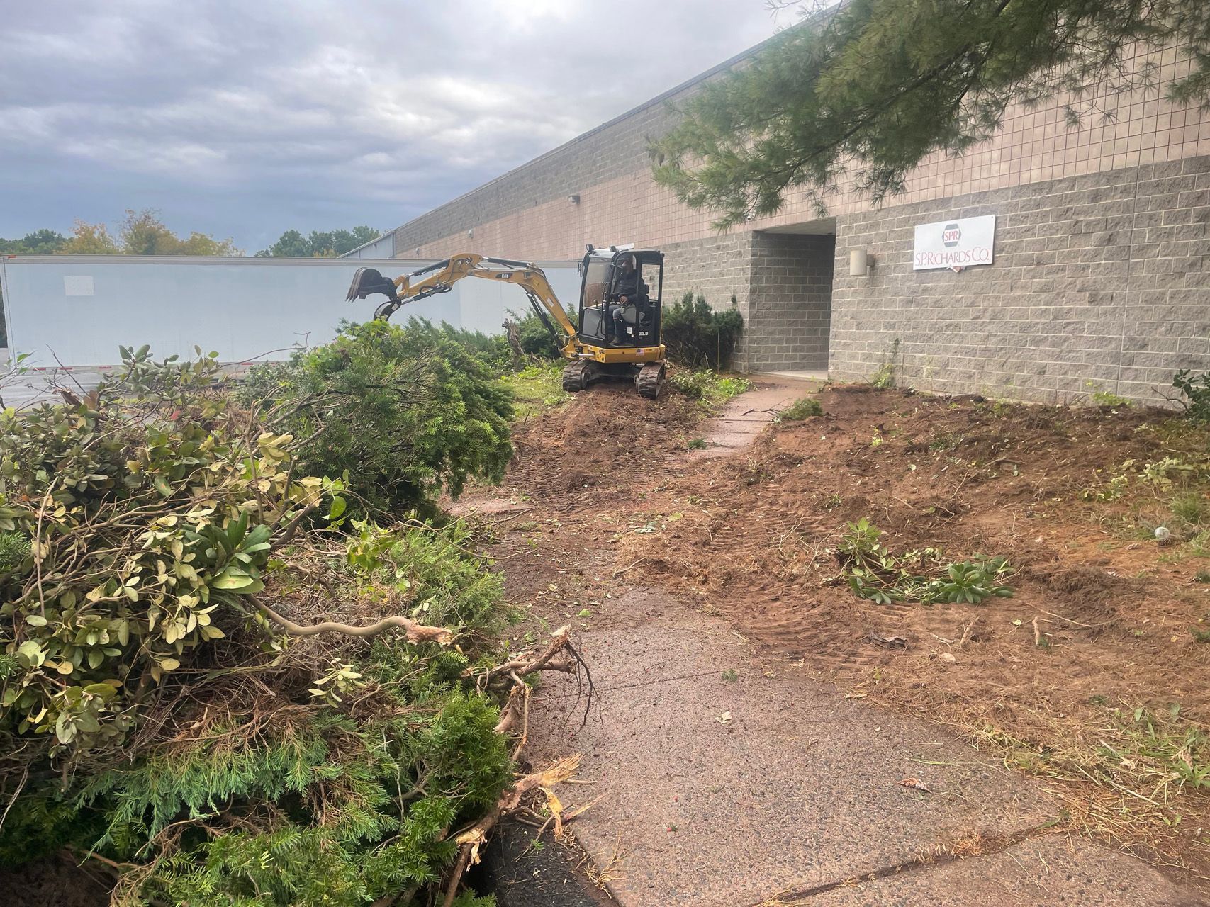 Small excavator clearing brush beside a concrete building and path on a cloudy day