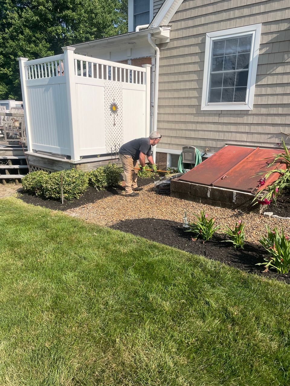 Person bending over in a front yard garden beside a house and white deck railing