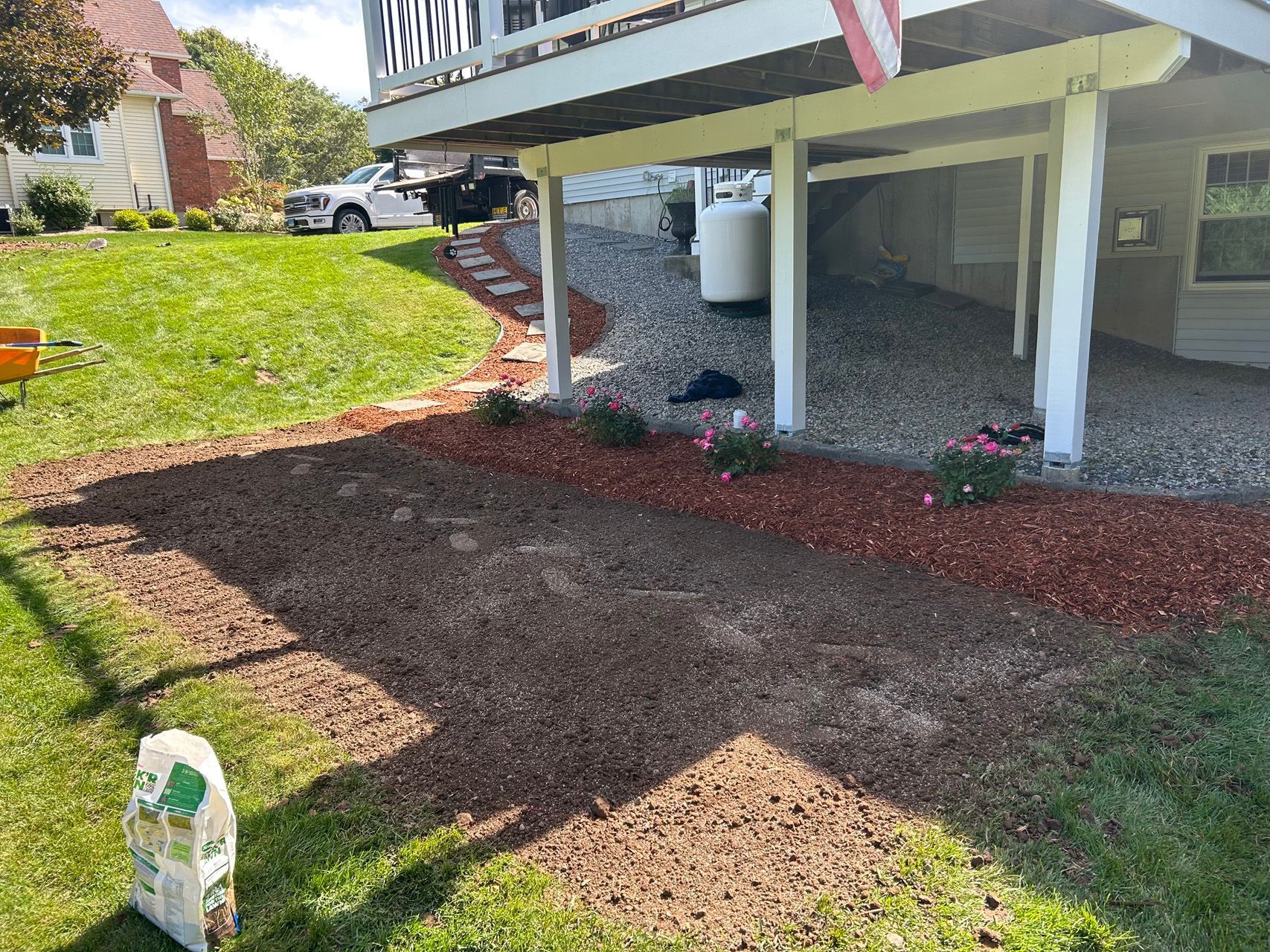 Mulched garden bed beside a raised porch with white posts and a grassy slope in sunlight