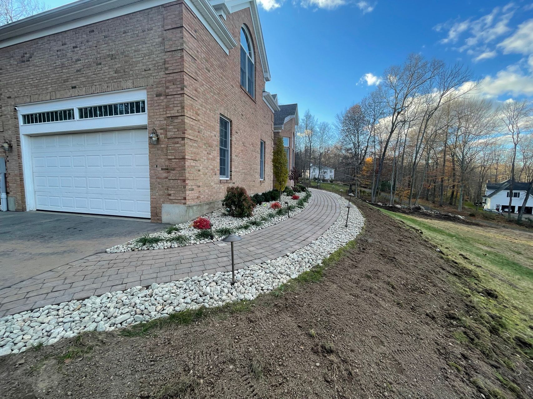 Brick building with white garage door, curved stone walkway, and landscaped lawn beside wooded area.