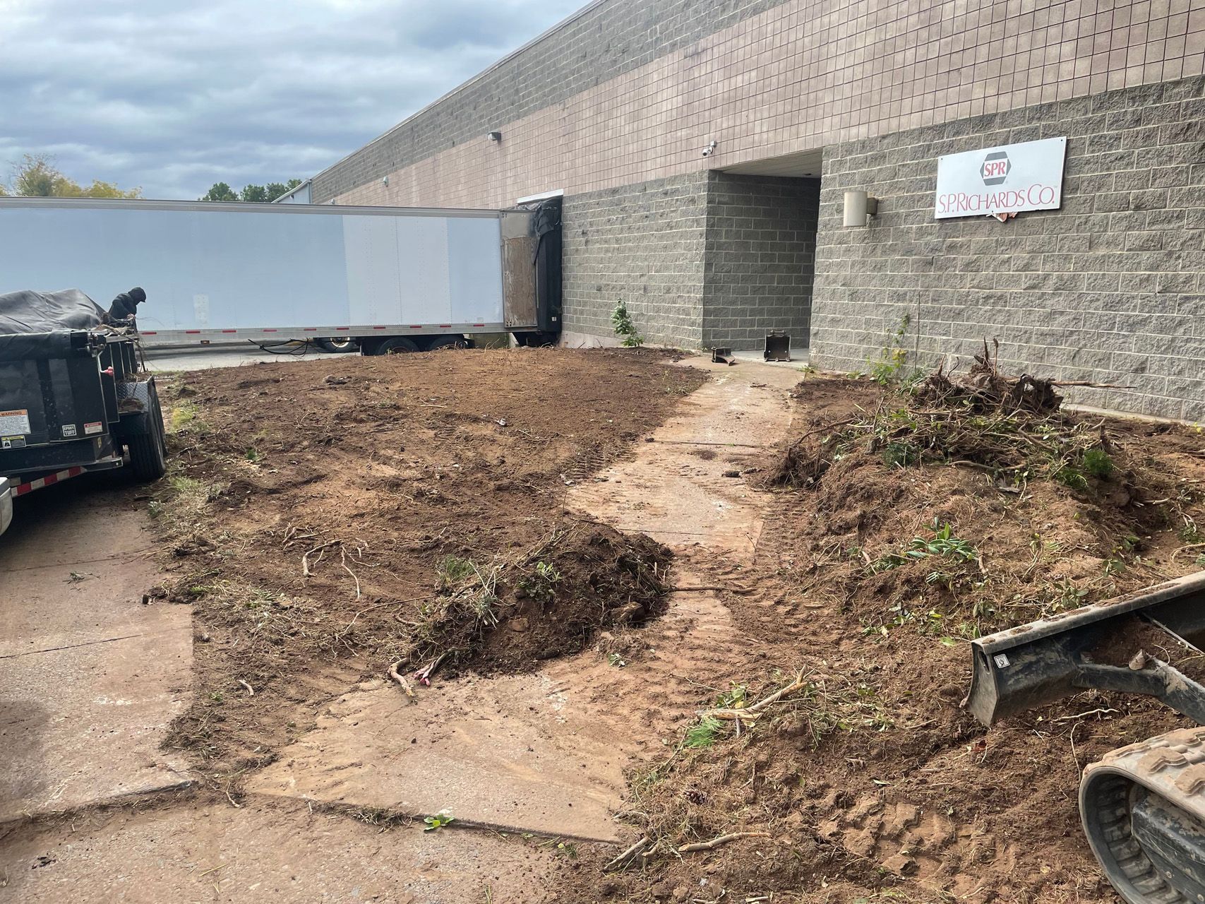 Dirt-covered construction site beside a brick building with a trailer and scattered equipment.