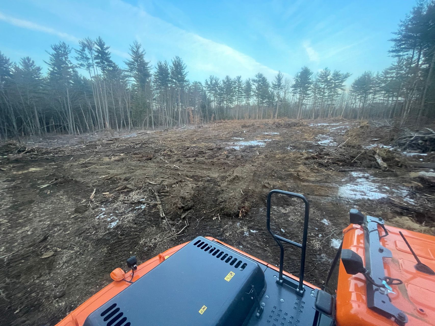 Orange tractor cab overlooking a muddy, cleared woodland area with snow patches and trees in the distance