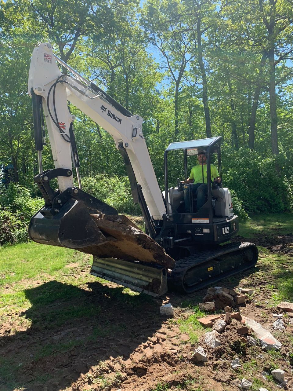 White excavator digging soil in a wooded area near a muddy trench