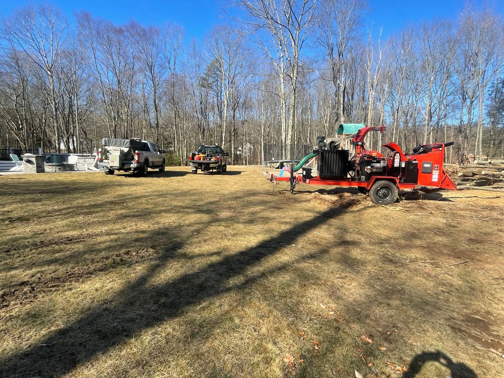 Red wood chipper in a grassy yard with two trucks and bare trees under a clear blue sky.