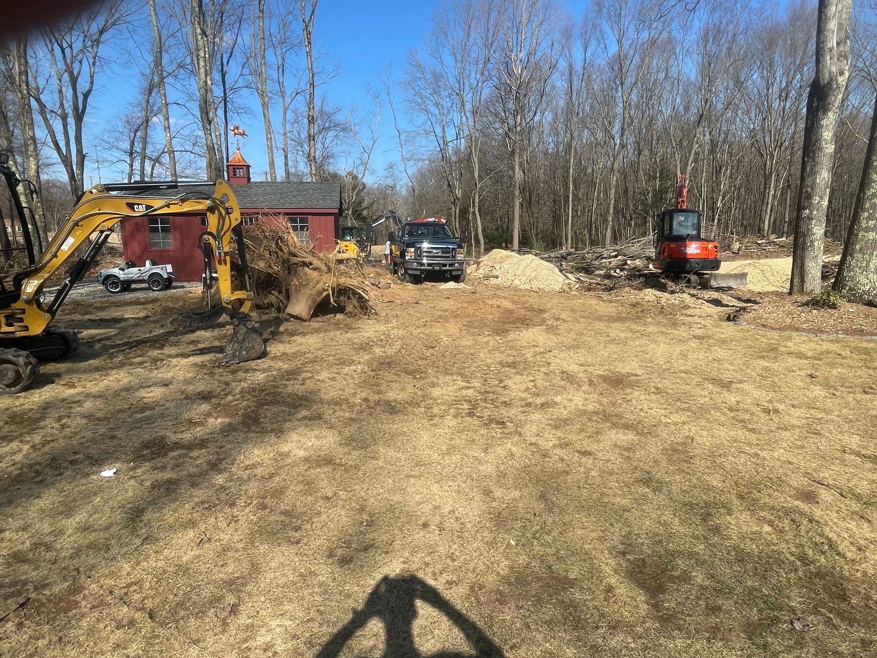 Construction equipment in a cleared wooded lot, with an excavator, dump truck, and small loader under a blue sky
